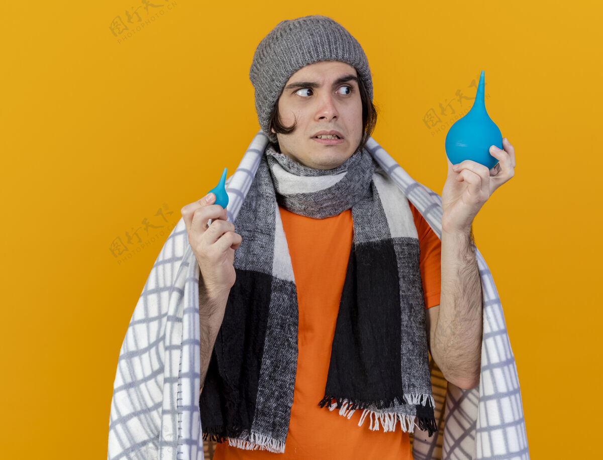 迷糊的困惑的年轻病人戴着冬天的帽子 围巾裹着格子布 手里拿着 看着隔离在橙色背景上的灌肠围巾帽子包着的