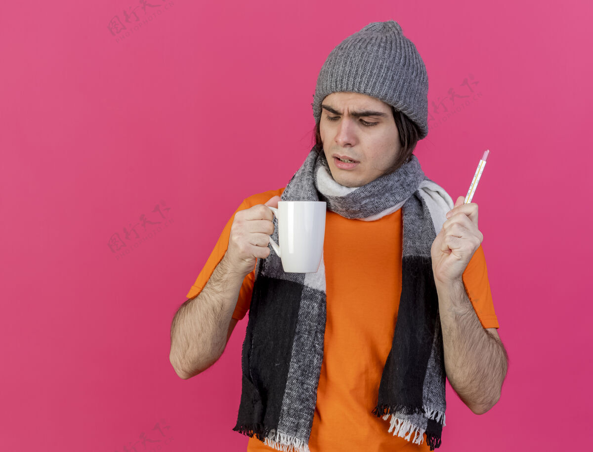 体温计困惑的年轻病人戴着冬天的帽子 戴着围巾 拿着温度计 看着手中的一杯茶 孤立在粉红色的背景上抱着帽子生病