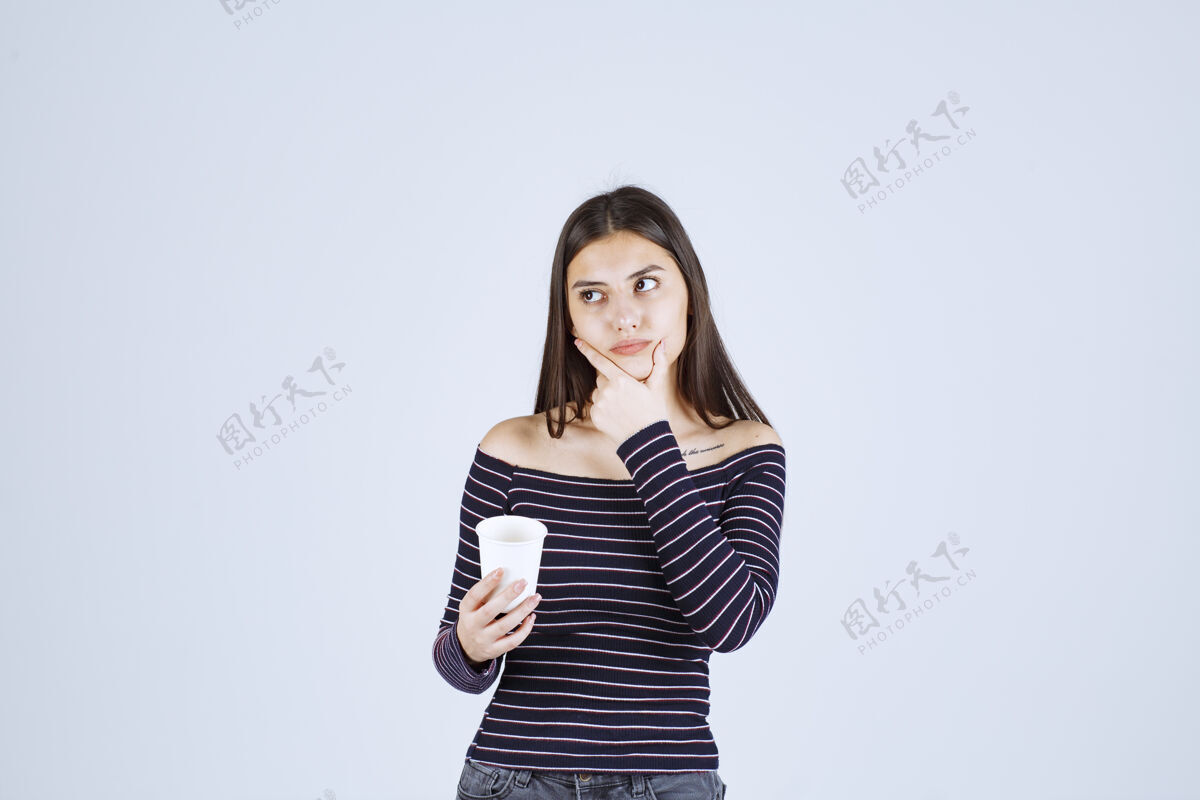 品味穿着条纹衬衫的女孩拿着一个塑料咖啡杯在思考雇员服装计划