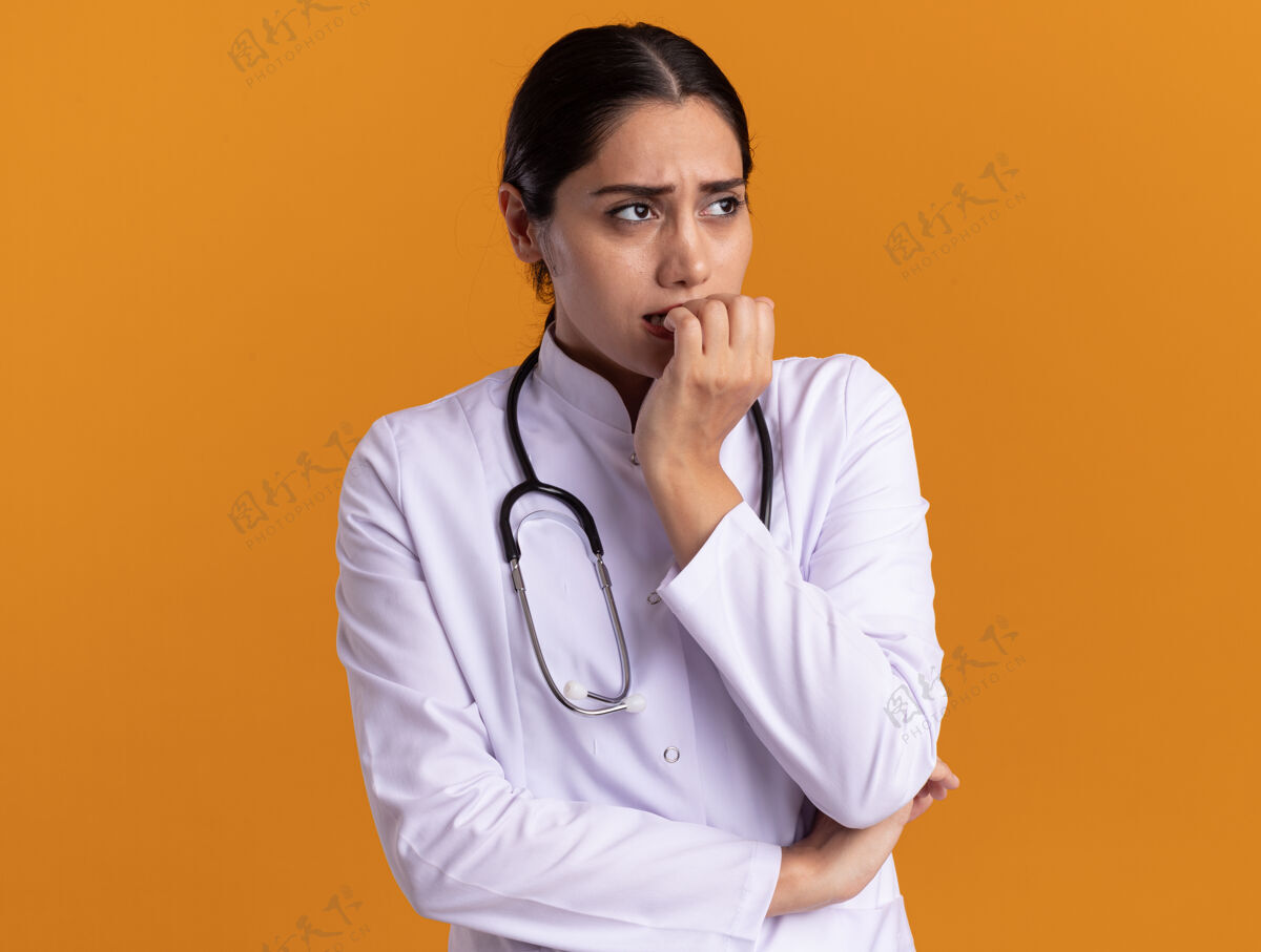 听诊器身穿医用外套的年轻女医生 脖子上戴着听诊器 站在橘色的墙上 紧张不安地看着旁边咬着的指甲女人外套压力