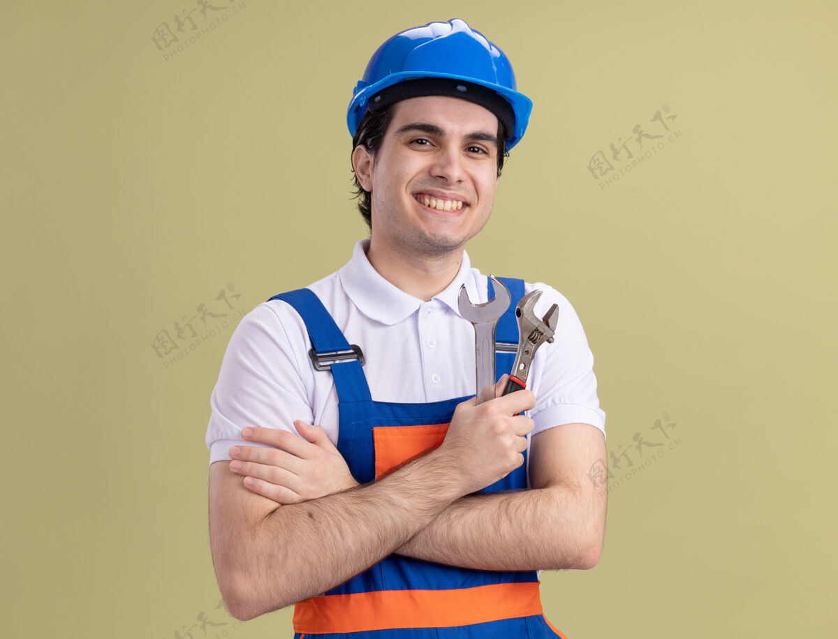 年轻人年轻的建筑工人穿着建筑制服 戴着安全帽 拿着扳手 面带微笑地站在绿色的墙上 望着前方安全建筑工人扳手