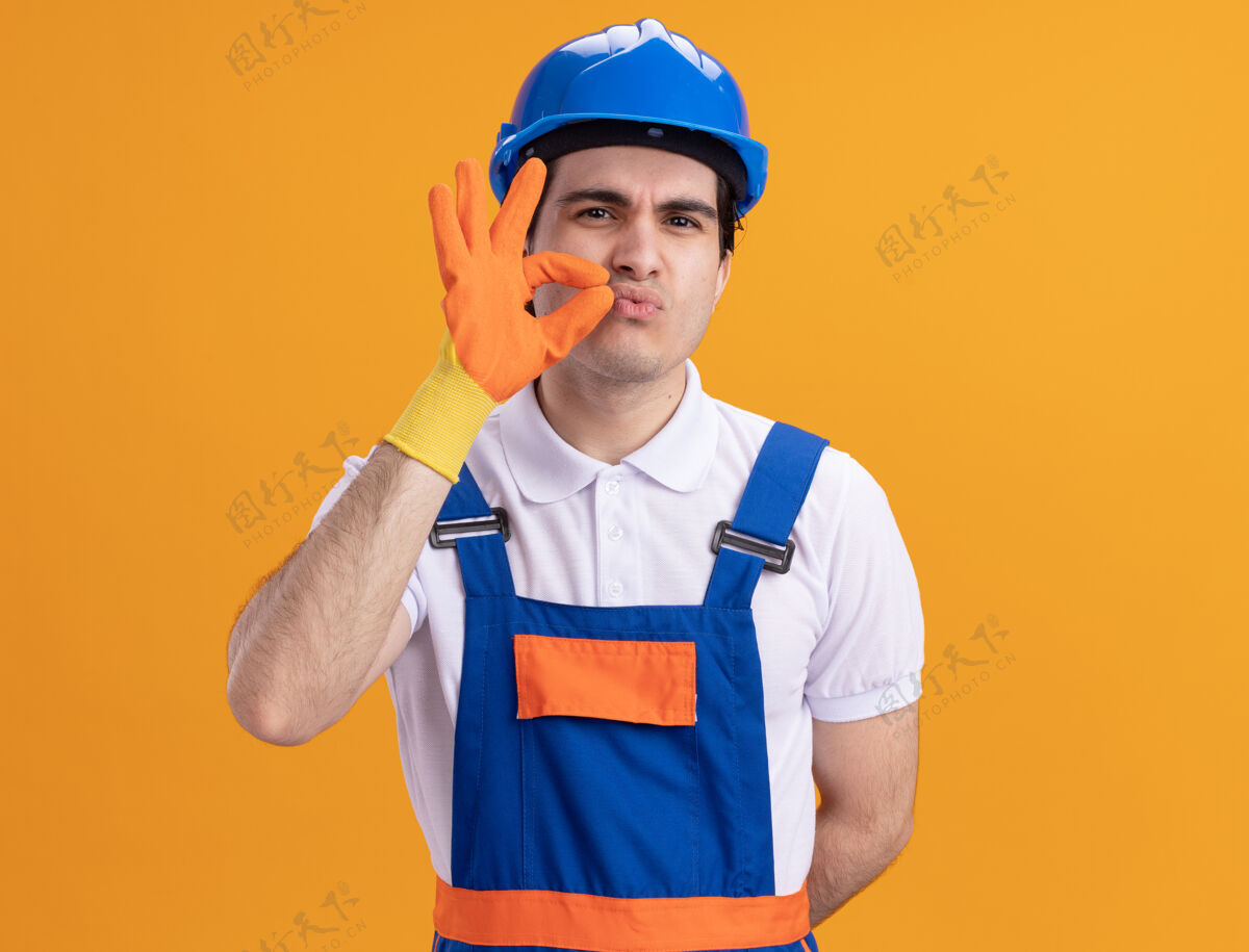 关闭愤怒的年轻建筑工人身穿建筑制服 戴着安全帽 戴着橡胶手套 站在橘色的墙上做着闭嘴的手势使喜欢建筑工人