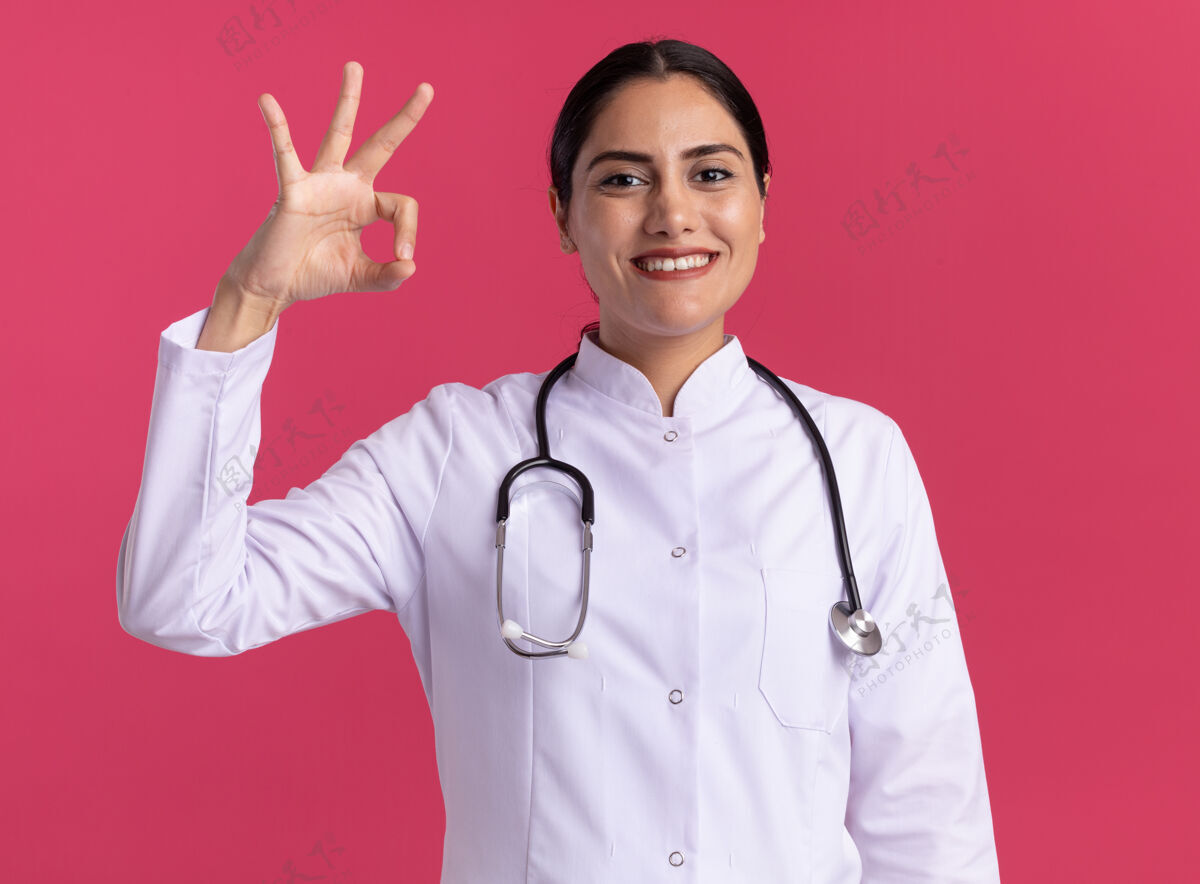 年轻身穿医用外套 手持听诊器的年轻女医生站在粉红色的墙上 面带微笑 脸上露出“ok”的标志医生开心医生