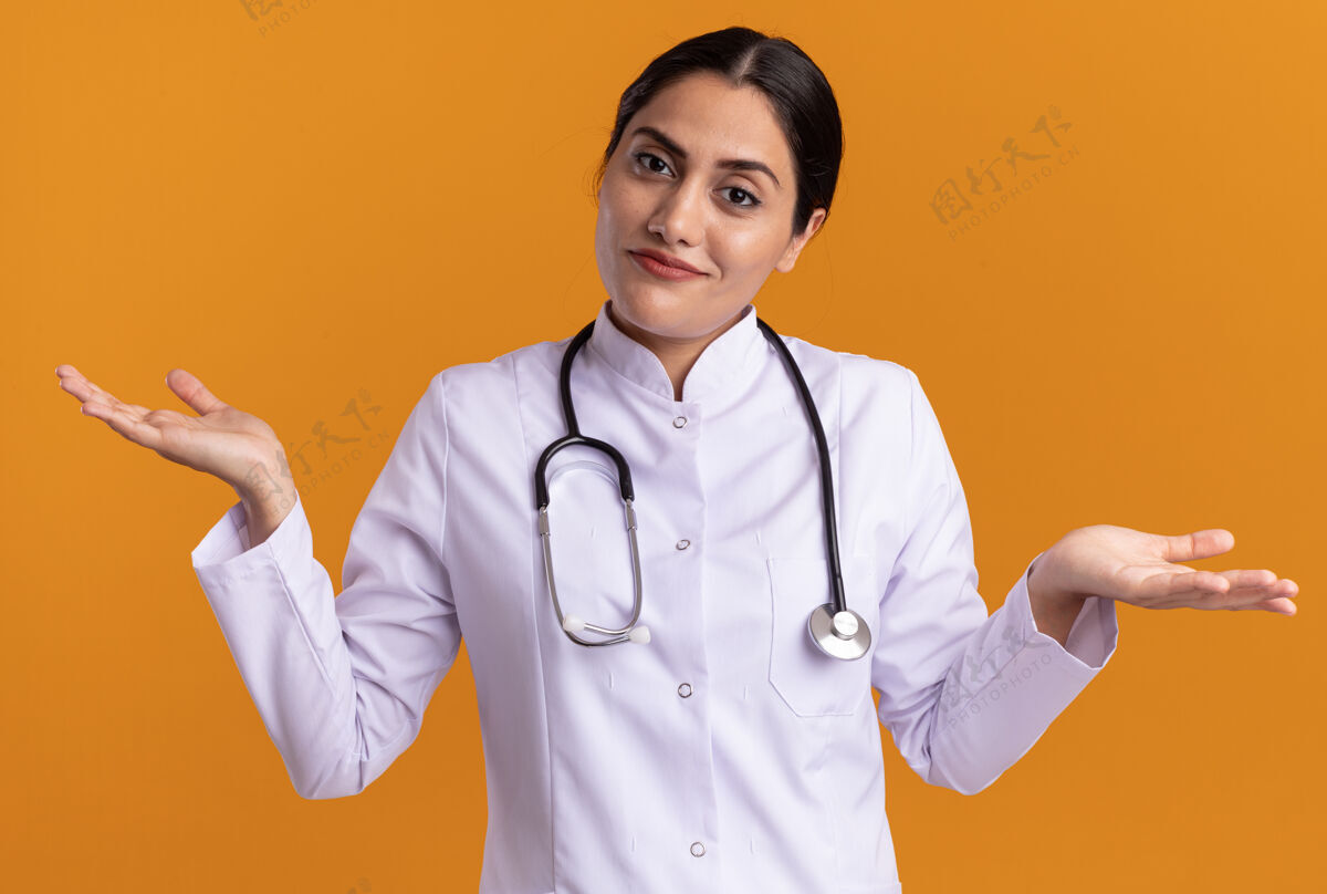 医生年轻的女医生 穿着医用外套 脖子上戴着听诊器 站在橘色的墙上 困惑地看着前面 两臂伸向两边 没有回答站外套困惑