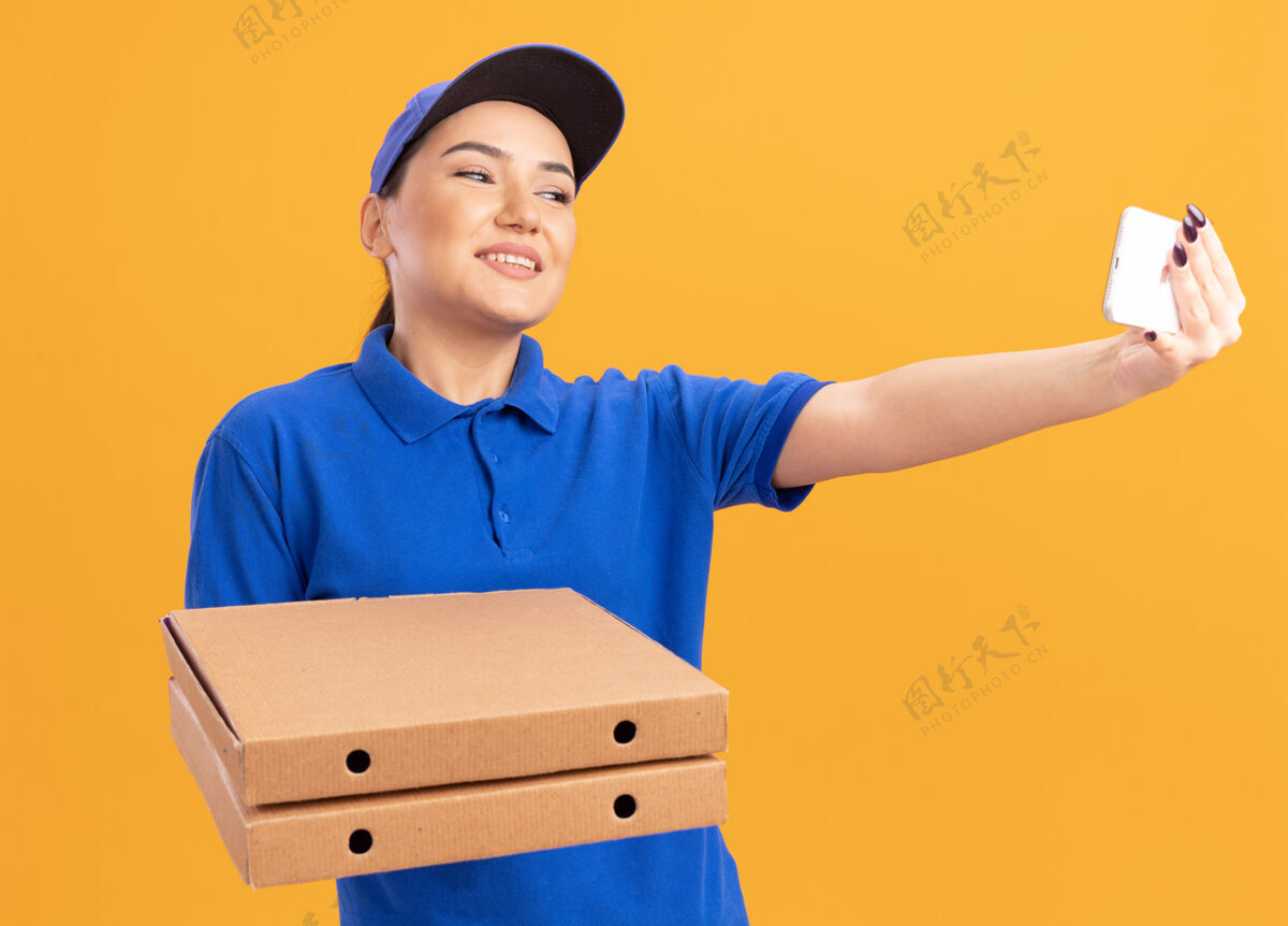 盒子身穿蓝色制服 戴着帽子 拿着披萨盒 用智能手机自拍的年轻女送货员站在橙色的墙上开心地微笑着自拍女人站着