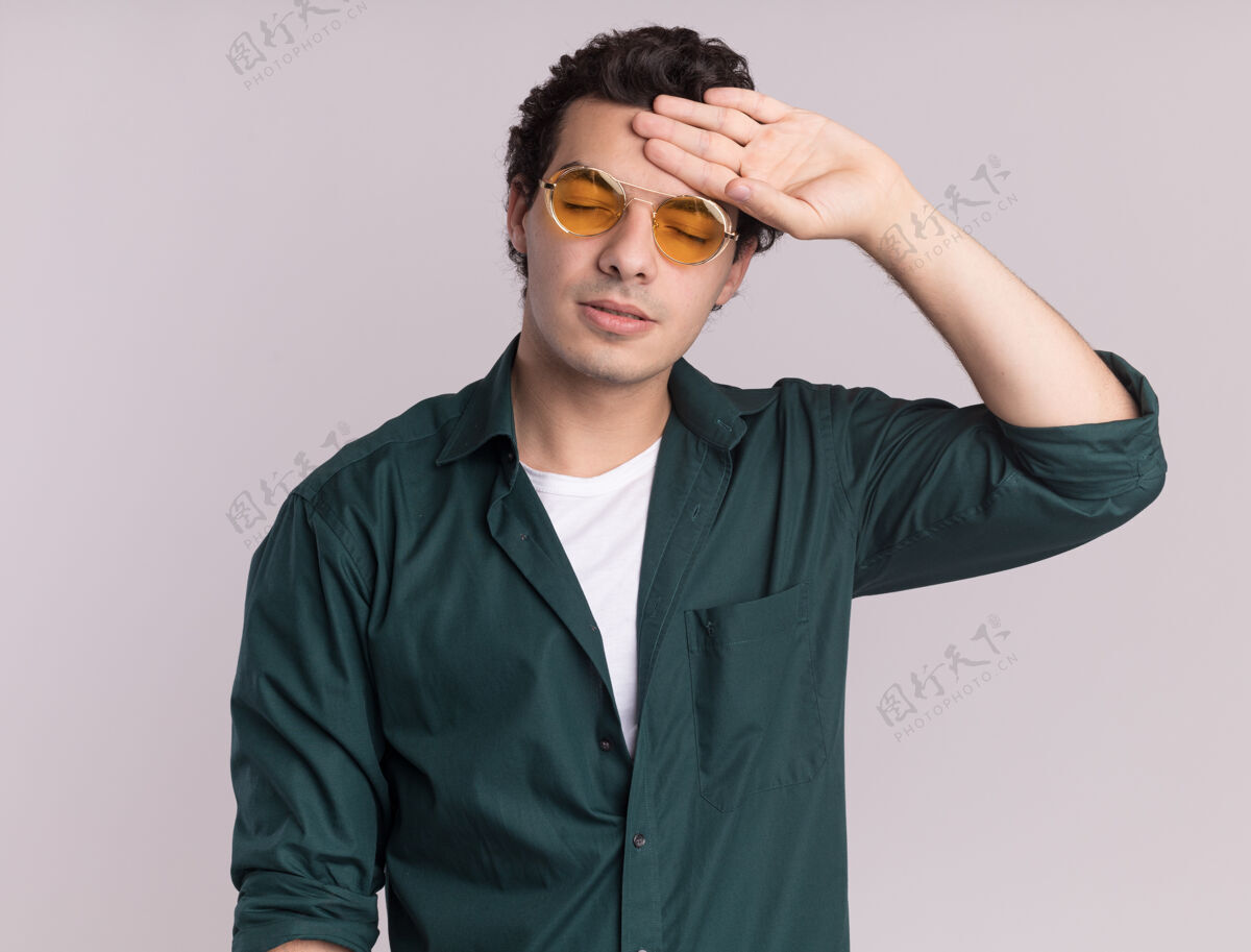 年轻一个穿着绿衬衫戴着眼镜的年轻人站在白墙上 看上去很累 很无聊男人无聊额头