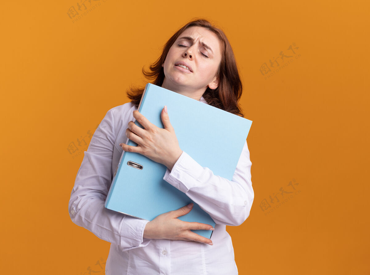 年轻身穿白衬衫的年轻女子站在橙色的墙上 手里拿着文件夹 看上去很疲惫 而且工作过度过度劳累站着女人