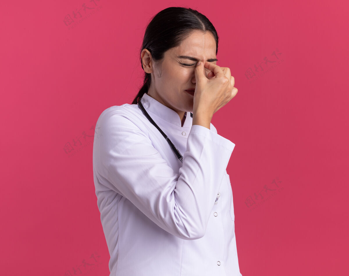 年轻人穿着医用外套的年轻女医生站在粉红色的墙上 用听诊器在闭着的眼睛之间触摸鼻子 脸上露出恼怒的表情外套闭着眼睛