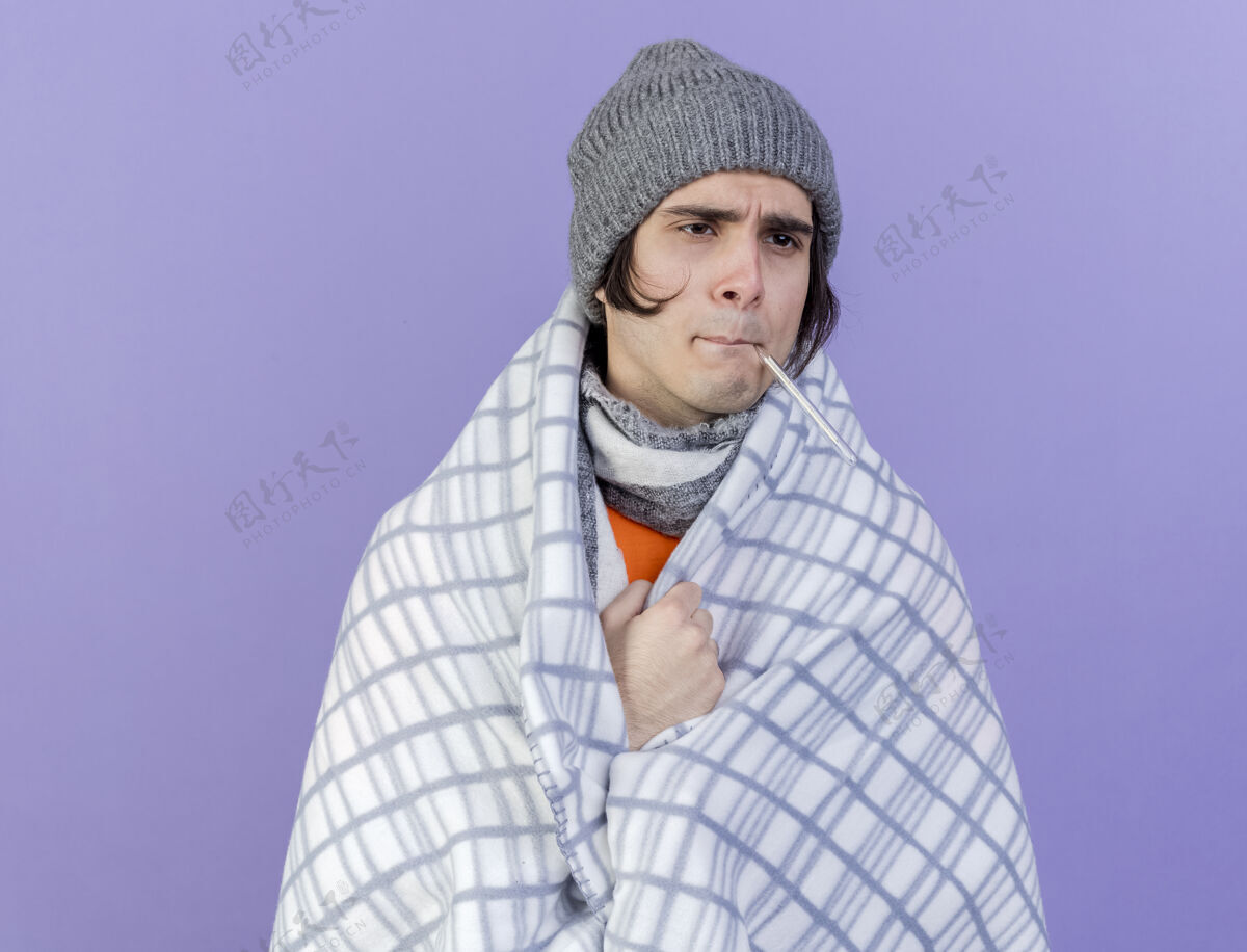 嘴望着身边体弱多病的年轻人戴着冬天的帽子 披着方格围巾 嘴里叼着温度计 隔离在紫色的背景上抱疾病格子布