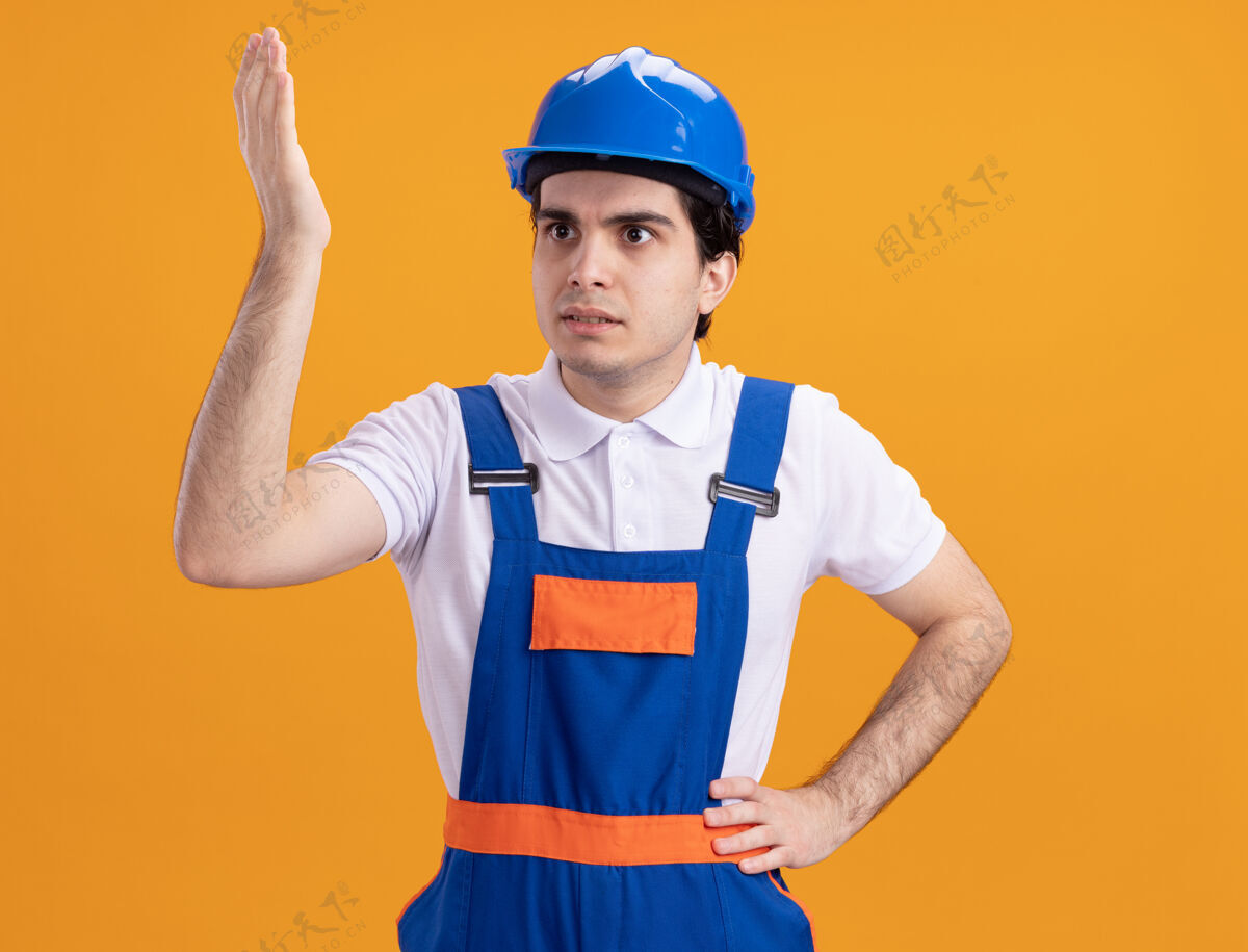 建设者身穿建筑制服 戴着安全帽的年轻建筑工人站在橙色的墙上 困惑地向一旁望去 不高兴地举起手来建筑困惑年轻