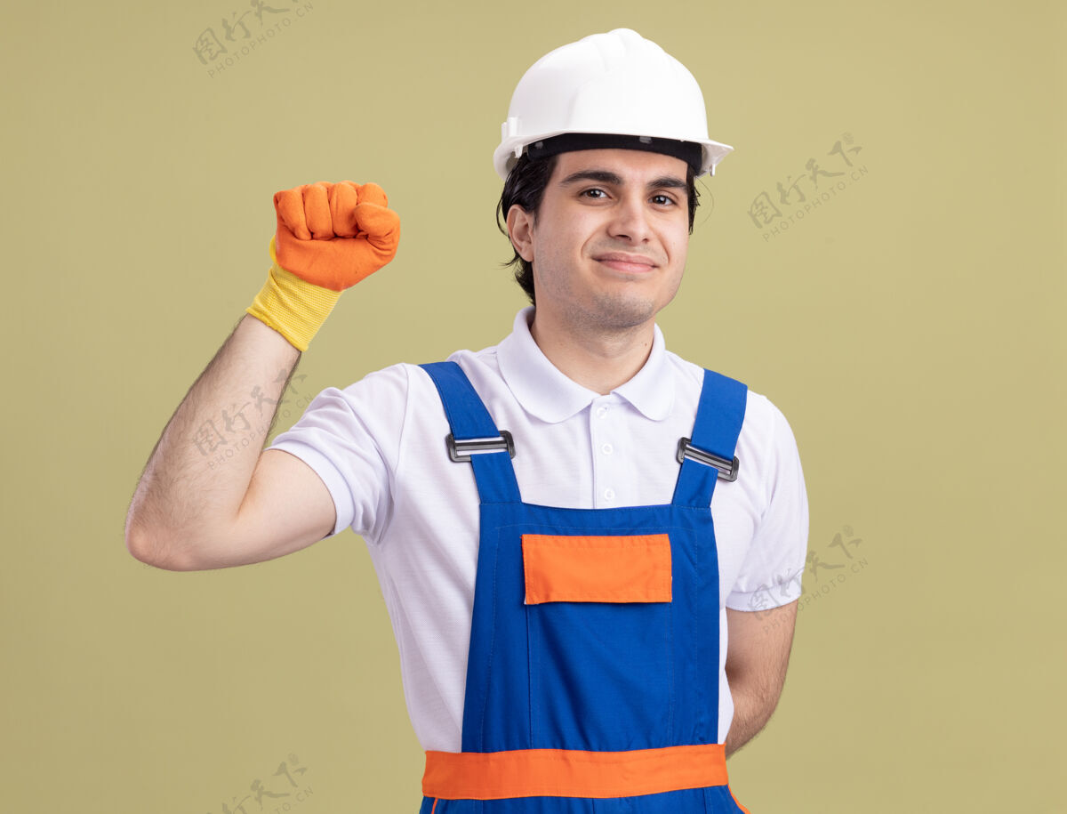 信心年轻的建筑工人穿着建筑制服 戴着橡胶手套戴着安全帽 微笑着自信地举起拳头站在绿色的墙壁上看着前方安全头盔提高