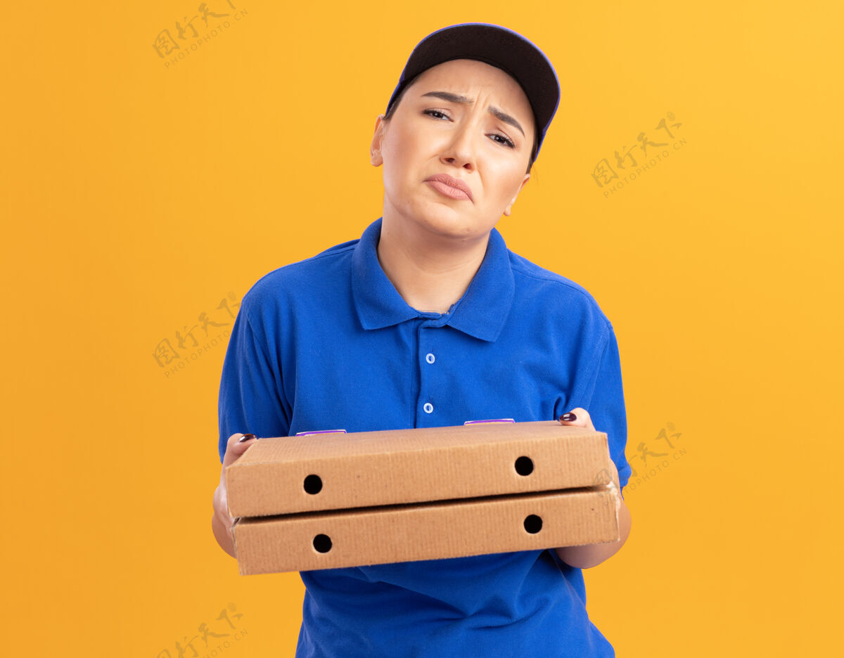 悲伤身穿蓝色制服 戴着帽子 拿着披萨盒 站在橘色的墙上 愁眉苦脸地看着前方的不幸的年轻送货员女人盒子帽子