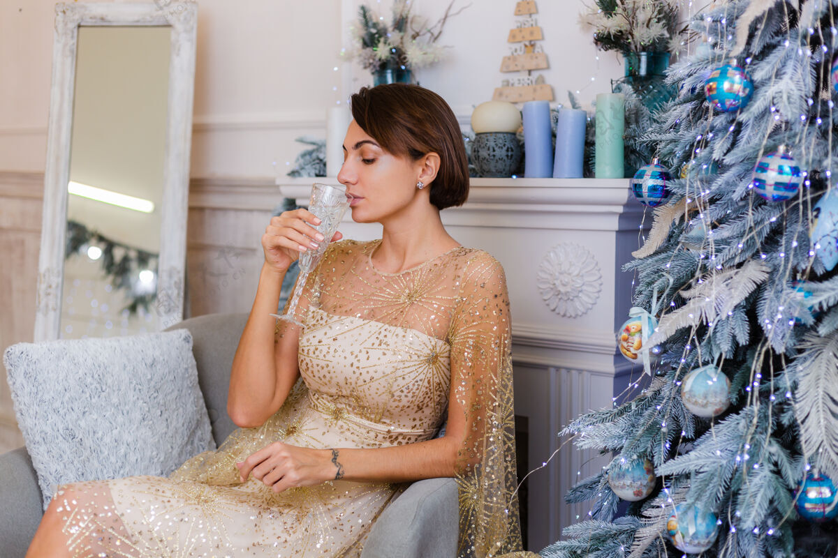 性感一个穿着闪亮的金色圣诞晚礼服的女人在家里拿着一杯香槟在圣诞树旁圣诞节房子吐司