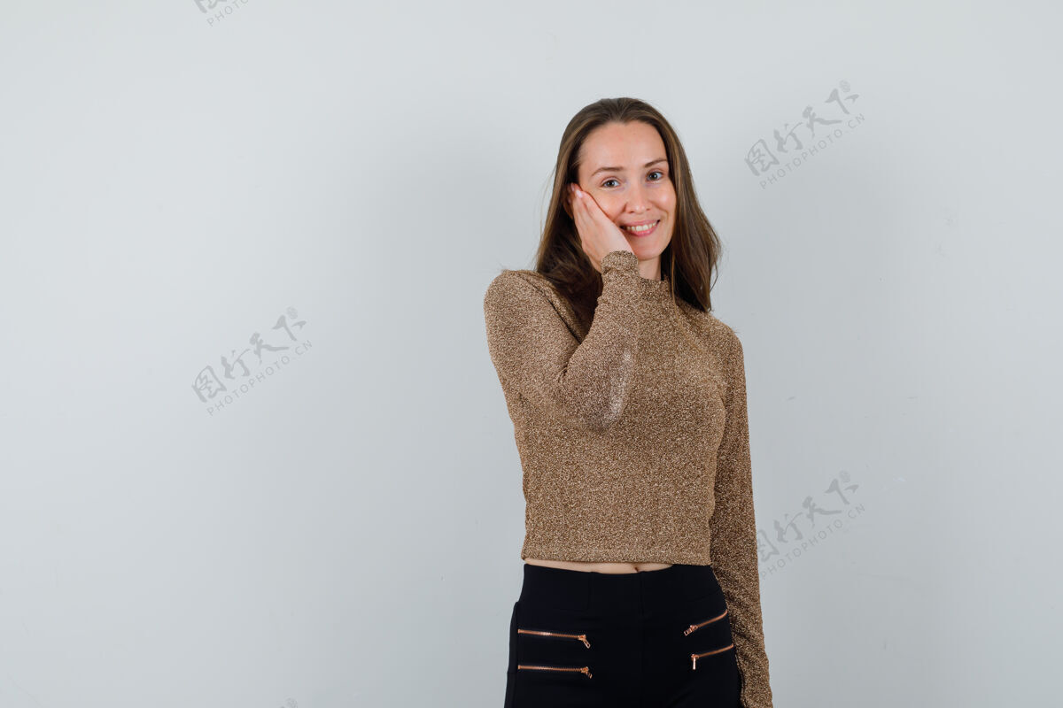 头发年轻女子面颊靠在手掌上 穿着金色的毛衣和黑色的裤子 看起来很幸福前面模特成人