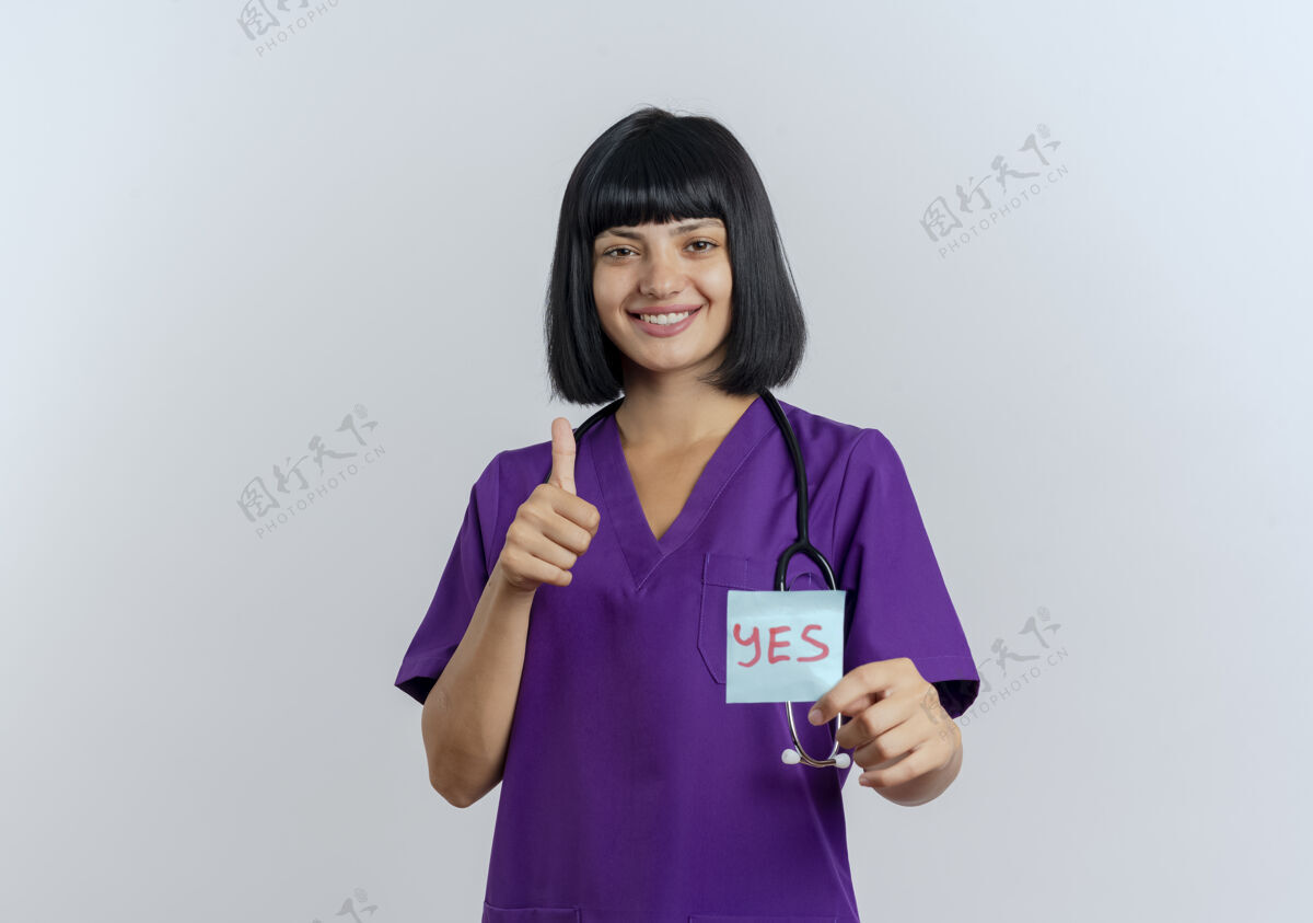 医生微笑着的年轻黑发女医生穿着制服 手持听诊器 竖起大拇指 手里拿着“是”字条拇指笔记制服