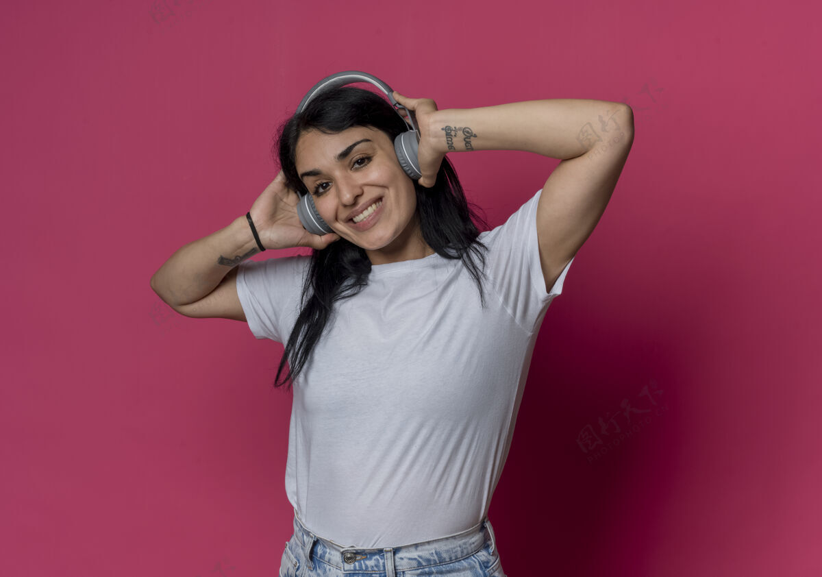 年轻微笑的年轻黑发白人女孩手持耳机孤立在粉红色的墙上粉色女人耳机