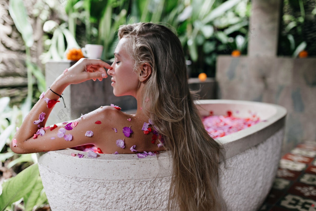 享受一个留着长直发的女人坐在满是玫瑰花瓣的浴缸里室内拍摄了一个漂亮的晒黑女人在家休息和做spa金发欧洲休闲