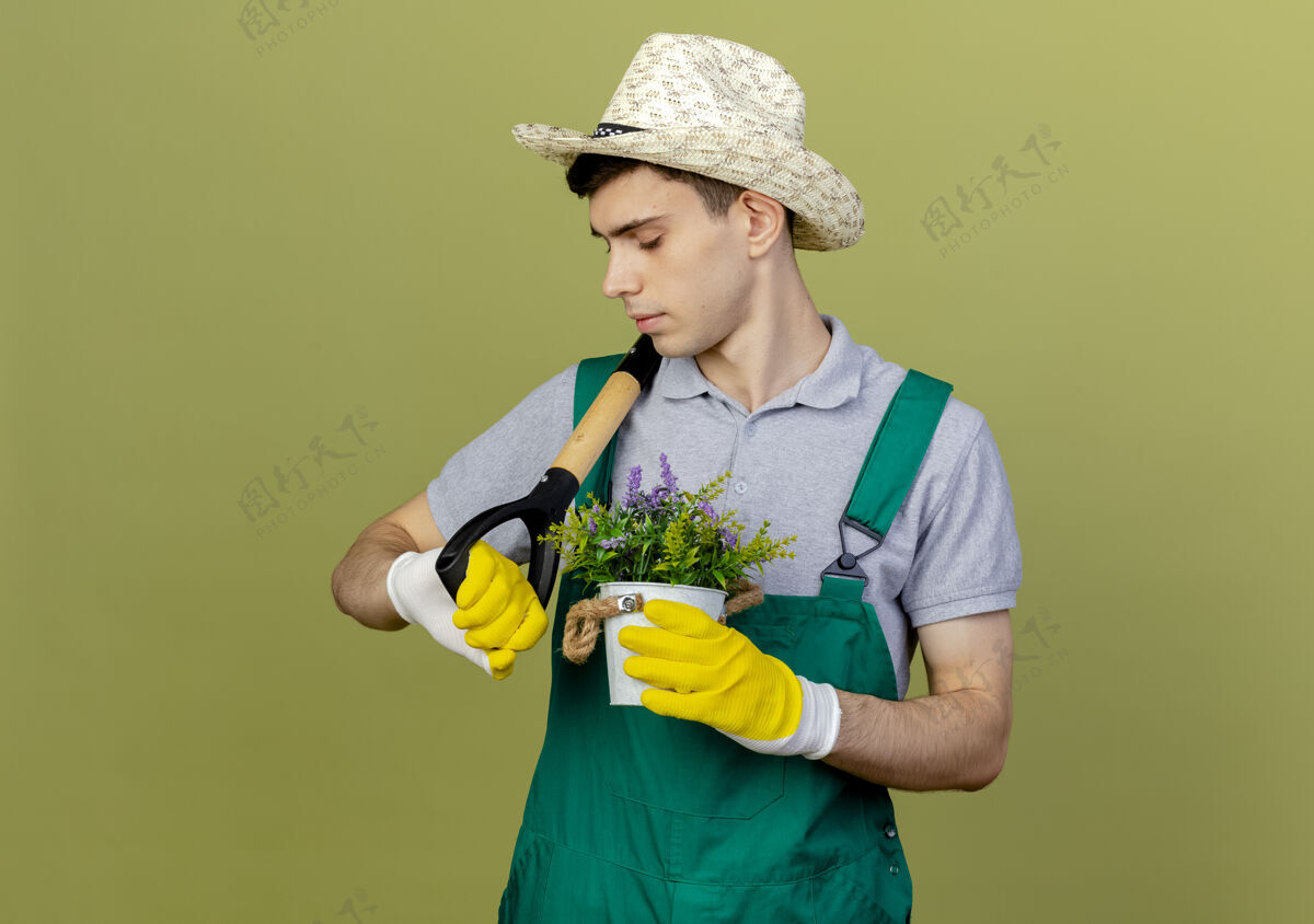 黑桃自信的年轻男园丁戴着园艺帽和手套 肩上扛着铁锹 花盆里放着鲜花绿色花空间