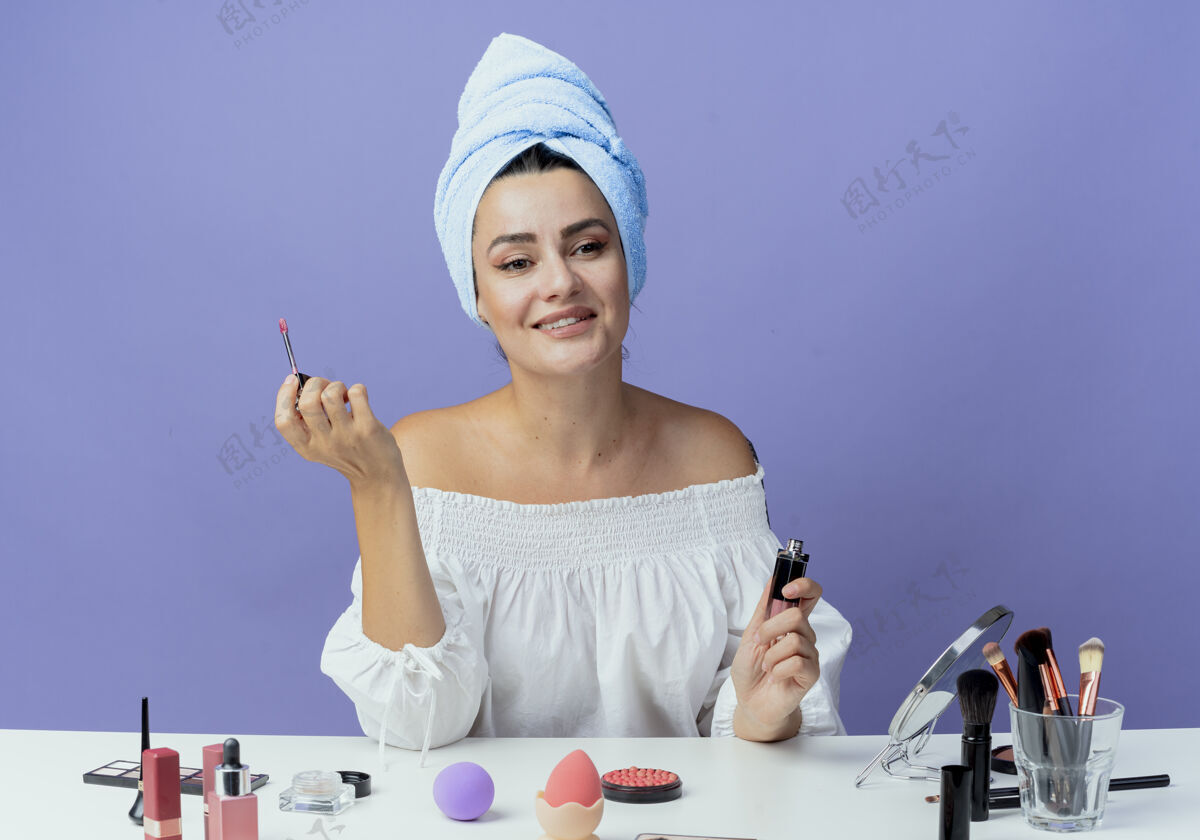 光泽微笑美丽的女孩裹着发巾坐在桌子上 用化妆工具夹着唇彩 看着一边被隔离在紫色的墙上嘴唇坐着毛巾