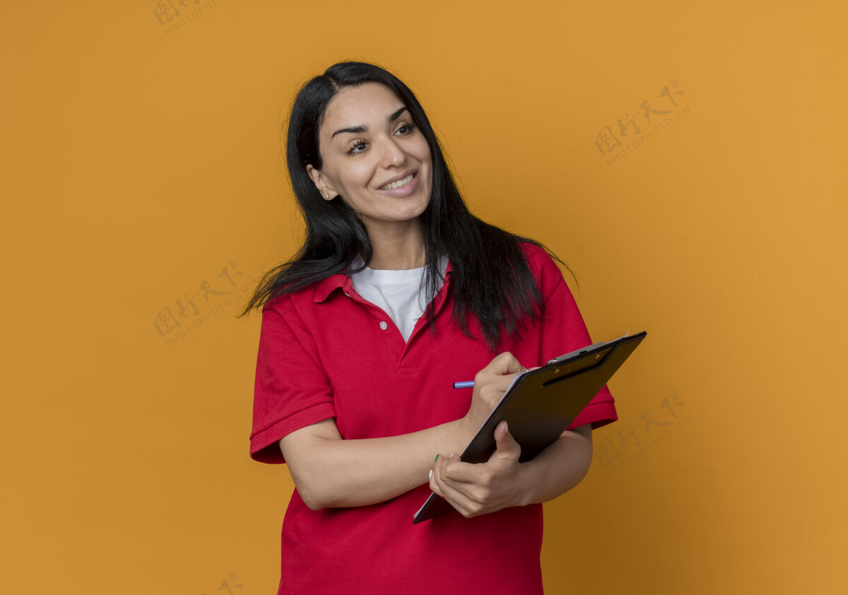 壁板微笑的年轻黑发白人女孩穿着红色衬衫拿着笔和剪贴板看着橙色墙上孤立的一面钢笔红色微笑