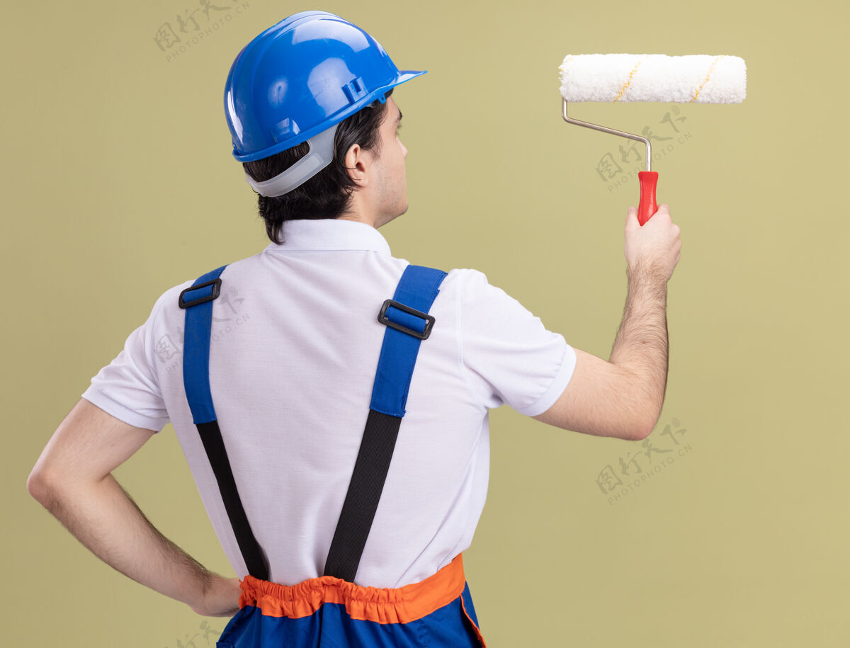 男人年轻的建筑工人穿着建筑制服 戴着安全帽 背着油漆辊站在绿色的墙上站着安全建筑工人