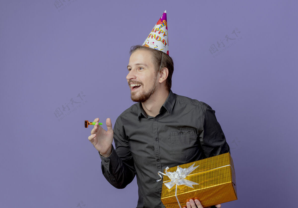 男人戴着生日帽的笑脸帅哥拿着礼盒吹着口哨看着紫色墙上孤立的一面生日帽子微笑
