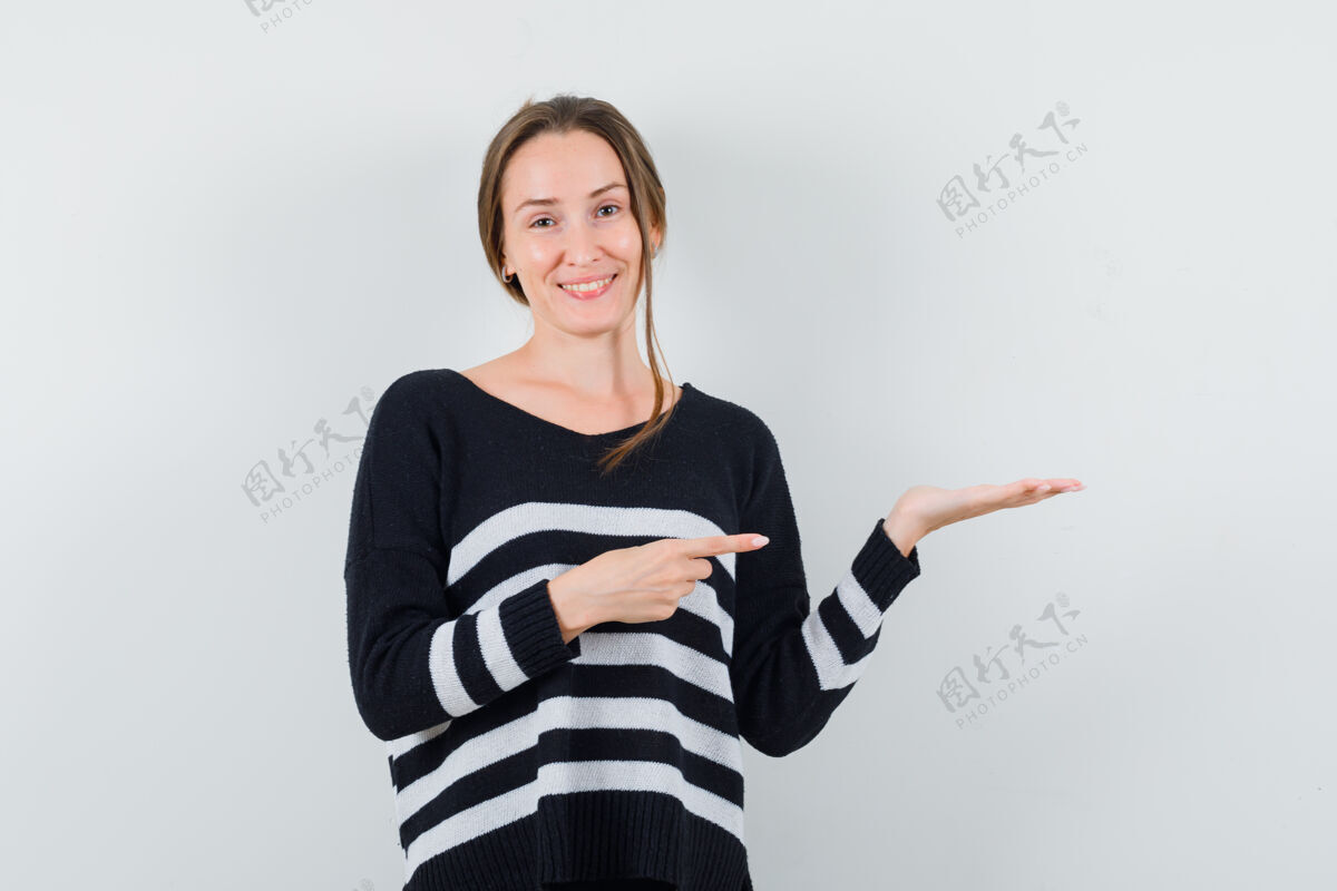 头发一个年轻的女人拿着一件虚构的东西 指着它 穿着条纹针织衫和黑色裤子 看起来很开心可爱持有指向