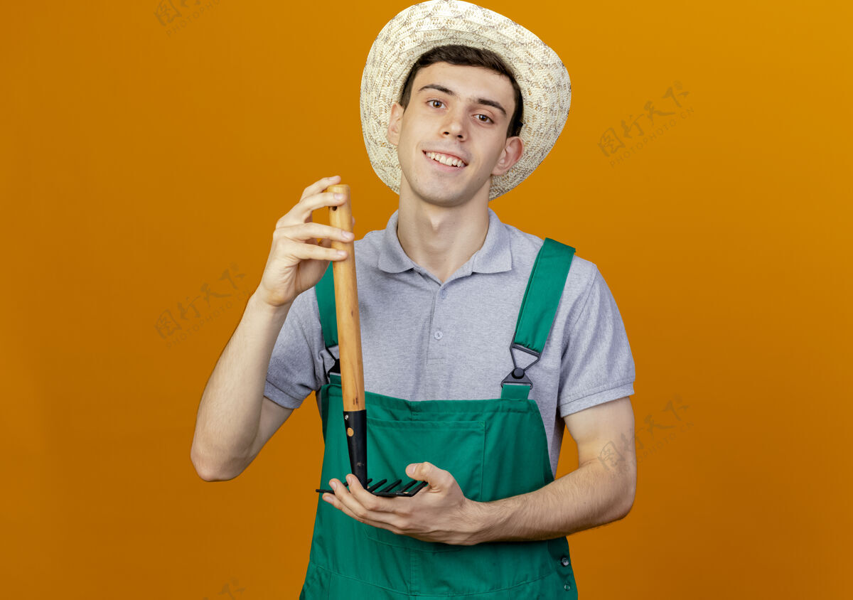 年轻微笑着的年轻男园丁戴着园艺帽 把耙子举得上下颠倒微笑花园帽子
