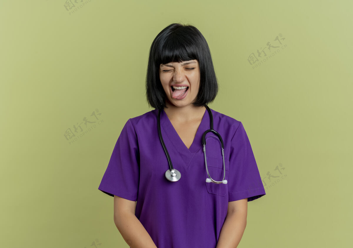 奥利弗恼怒的年轻黑发女医生穿着制服 手持听诊器 将舌头孤立在橄榄绿背景上 留有复印空间女性制服听诊器