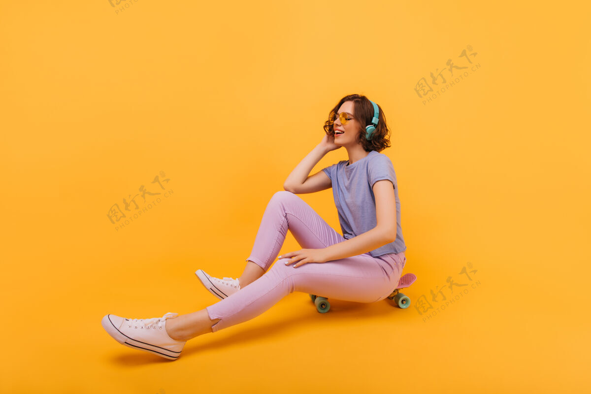 放松穿着时髦服装的快乐女人坐在滑板上闭着眼睛戴着黄色眼镜的漂亮女孩在长板上摆姿势休闲酷成人