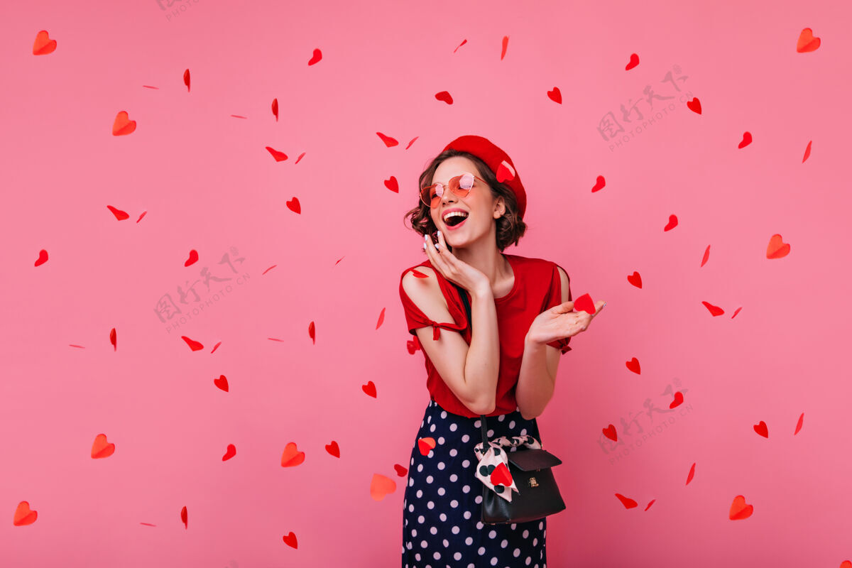 快乐时尚无忧无虑的女人在情人节摆姿势微笑迷人的卷发女孩在贝雷帽下站在红色的五彩纸屑女性表情欢呼