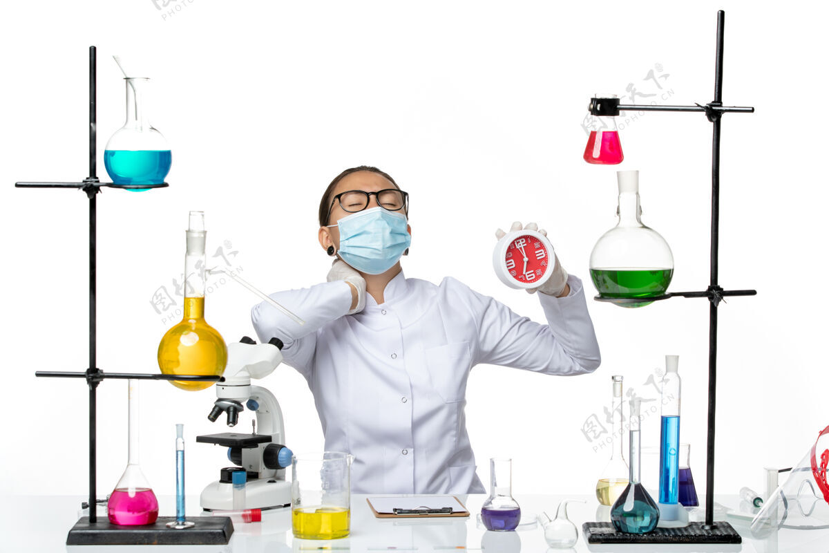 化学前视图：穿着医疗服的女化学家戴着面具拿着时钟 在白色背景上忍受着疼痛病毒实验室化学冠状病毒-飞溅持有实验室医学