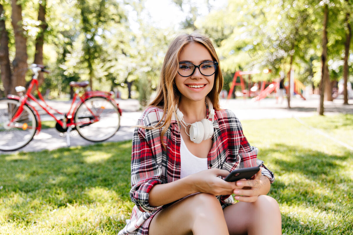 大笑可爱的欧洲女模特坐在草坪上打电话高加索美女在公园里和智能手机合影田野骑活动