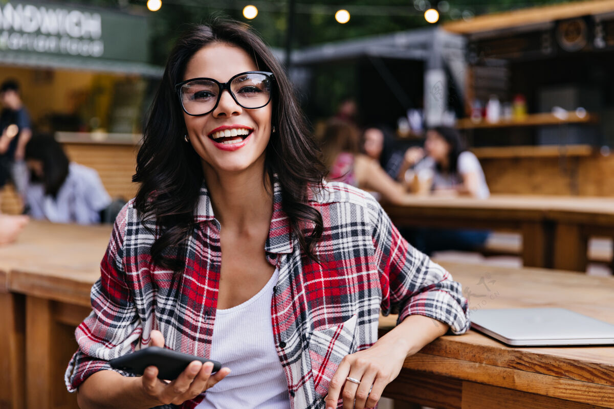 人时髦的白人女孩拿着智能手机微笑着在咖啡馆摆姿势美丽的女学生坐在户外餐厅与笔记本电脑自信笔记本电脑休闲