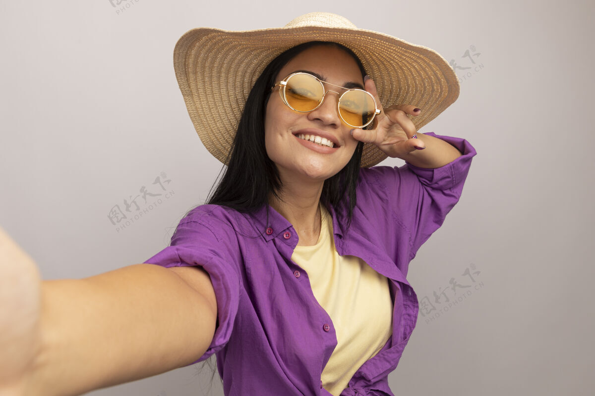 成人一个戴着太阳眼镜 头戴沙滩帽 脸上带着微笑的黑发白种人女孩 打着胜利手势 假装拿着相机在白色上自拍黑发海滩漂亮