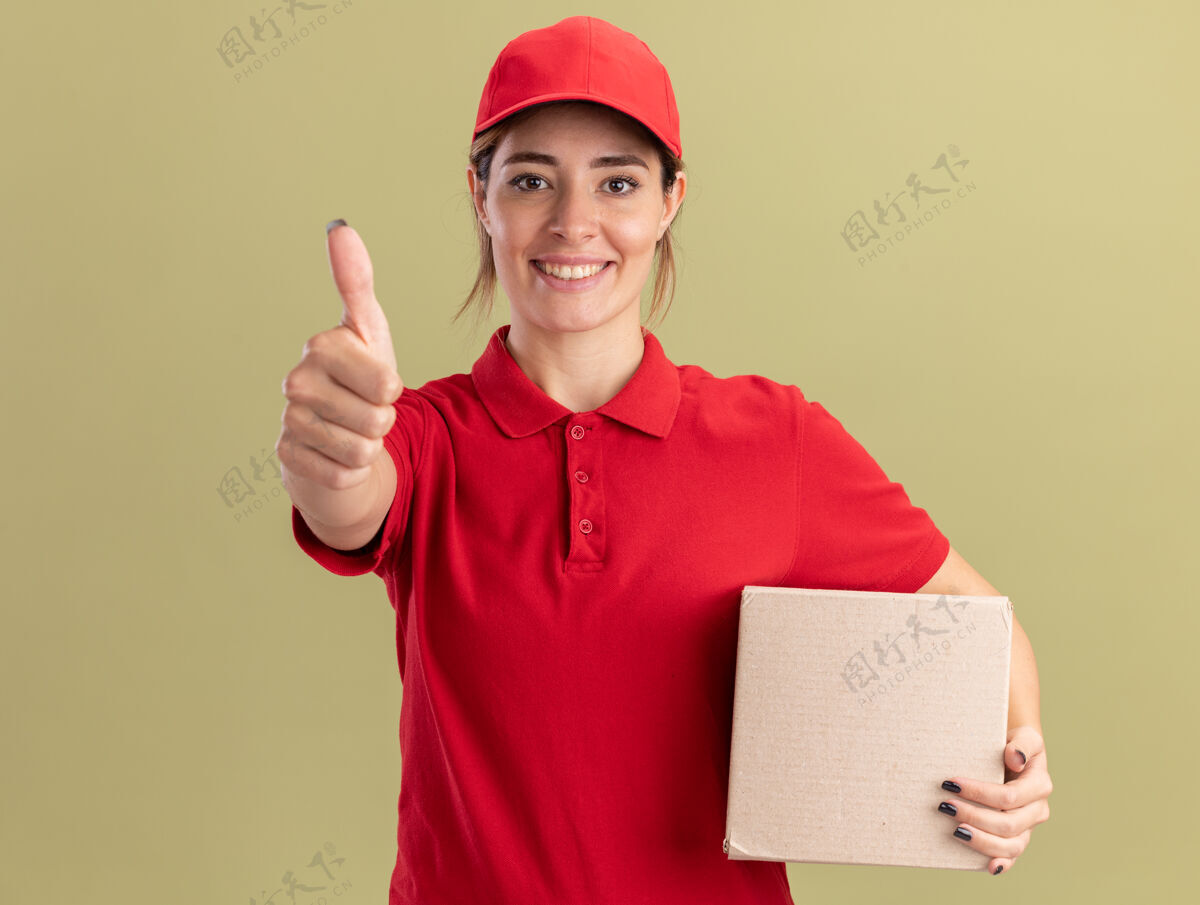 制服身着制服 面带微笑的年轻漂亮送货女孩竖起大拇指 拿着橄榄绿的名片盒橄榄漂亮姿势