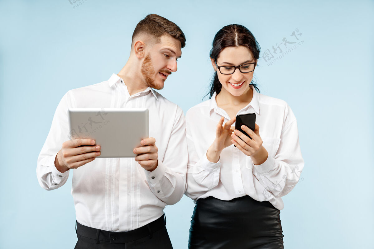 帅气商业伙伴关系的概念年轻快乐微笑的男女站在工作室蓝色背景下 手持手机和平板电脑团队合作在一起自信
