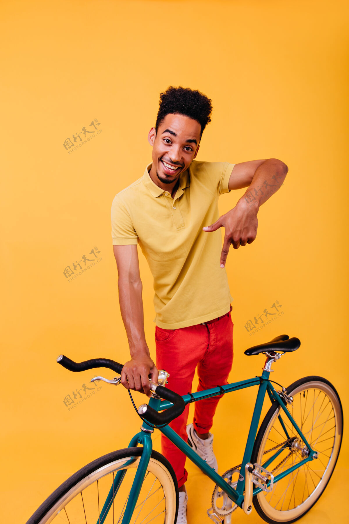 时尚一个穿着红裤子的笑脸黑人在玩的全长镜头一个骑自行车的活跃的非洲男人的室内肖像表情现代自行车