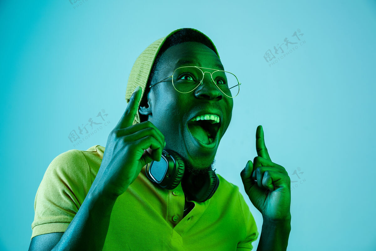 迪斯科年轻帅气的快乐惊喜的时髦男人在霓虹灯下用耳机听音乐迪斯科舞厅 夜总会 嘻哈风格 积极的情绪 面部表情 舞蹈概念帽子非洲音频