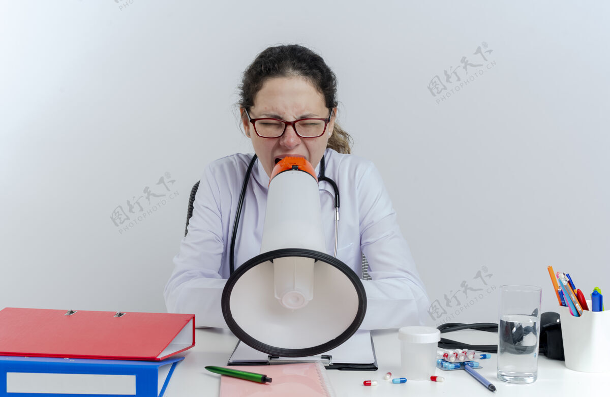 听诊器年轻的女医生穿着医用长袍 戴着听诊器和眼镜 坐在办公桌旁 拿着医疗工具 闭着眼睛大声喊叫年轻大声眼镜