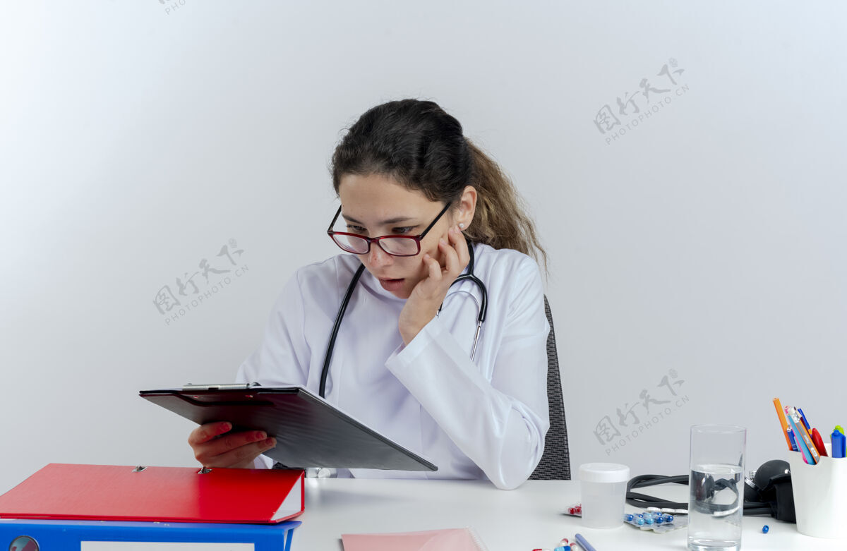 医疗年轻的女医生穿着医用长袍 戴着听诊器 戴着眼镜 坐在办公桌旁 拿着医疗工具 抚摸着脸 看着我女士工具成人