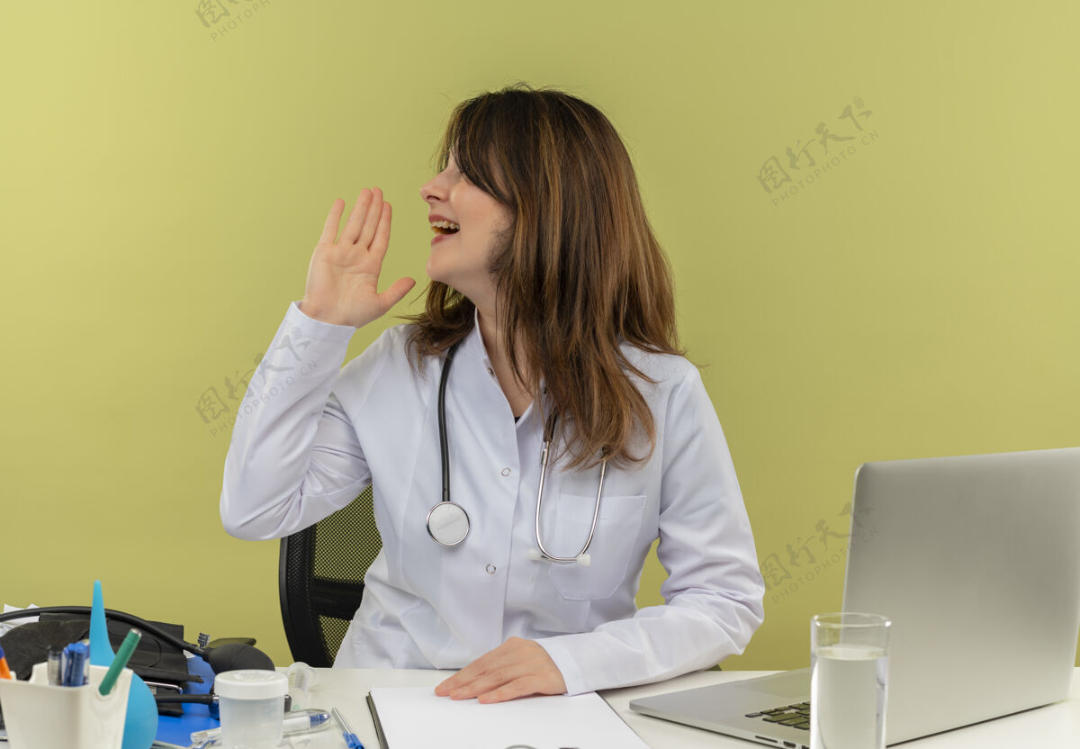医生令人印象深刻的中年女医生穿着医用长袍和听诊器坐在办公桌旁 手持医疗工具和笔记本电脑 头对侧地呼唤着一个孤独的人呼叫中年女士