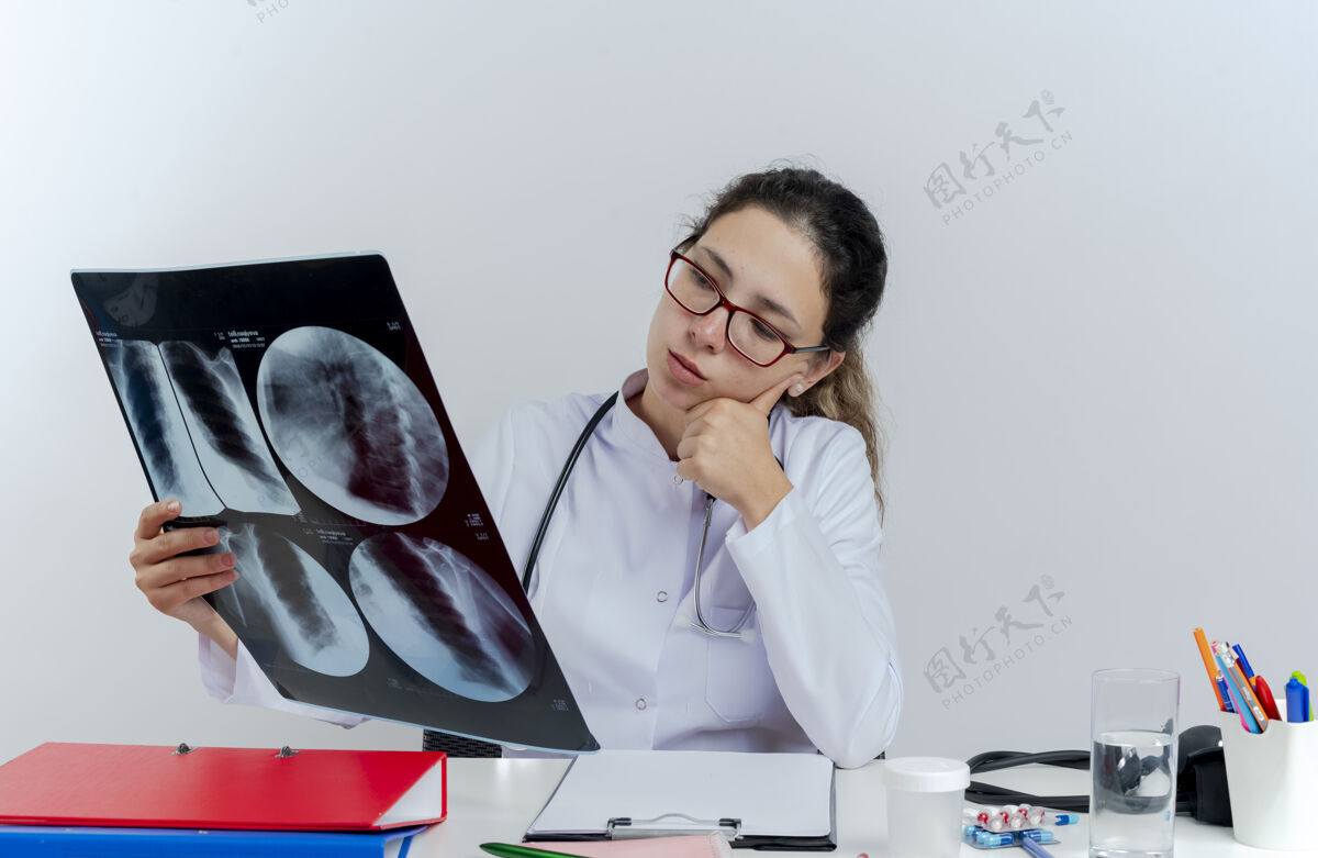 女人体贴的年轻女医生 穿着医用长袍 戴着听诊器 戴着眼镜 坐在办公桌旁 拿着医疗工具 看着x光片 手放在下巴上医疗长袍人