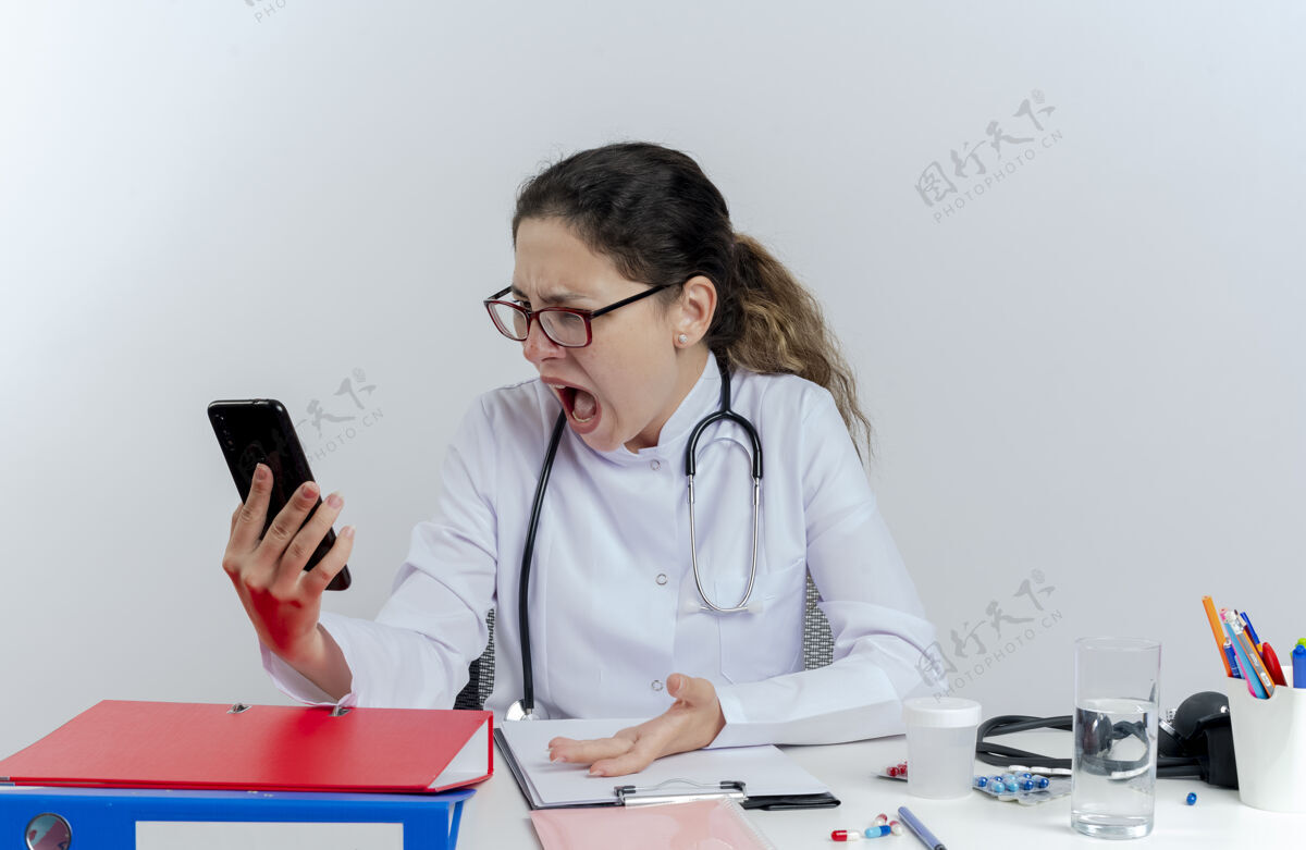 女性愤怒的年轻女医生穿着医用长袍 戴着听诊器和眼镜坐在办公桌旁 拿着医疗工具 孤立地看着手机女士听诊器电话
