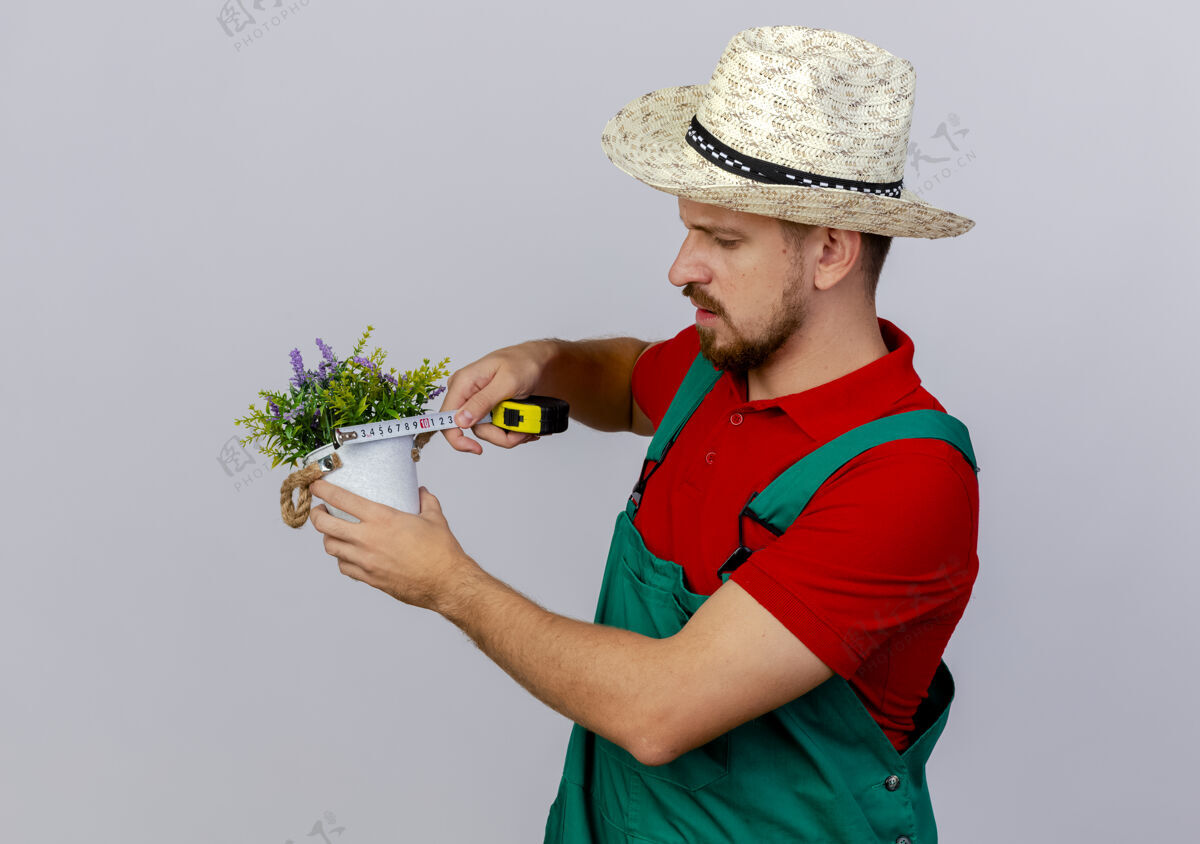 帽子年轻英俊的斯拉夫花匠穿着制服 戴着帽子 拿着花盆和卷尺测量它手测量带子