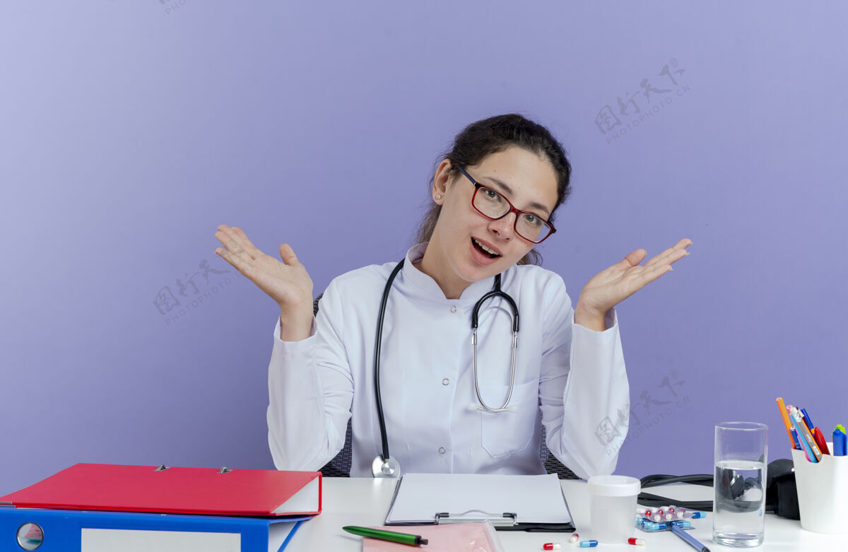 医生令人印象深刻的年轻女医生穿着医用长袍和听诊器坐在办公桌上与医疗工具看显示空手孤立紫色年轻人