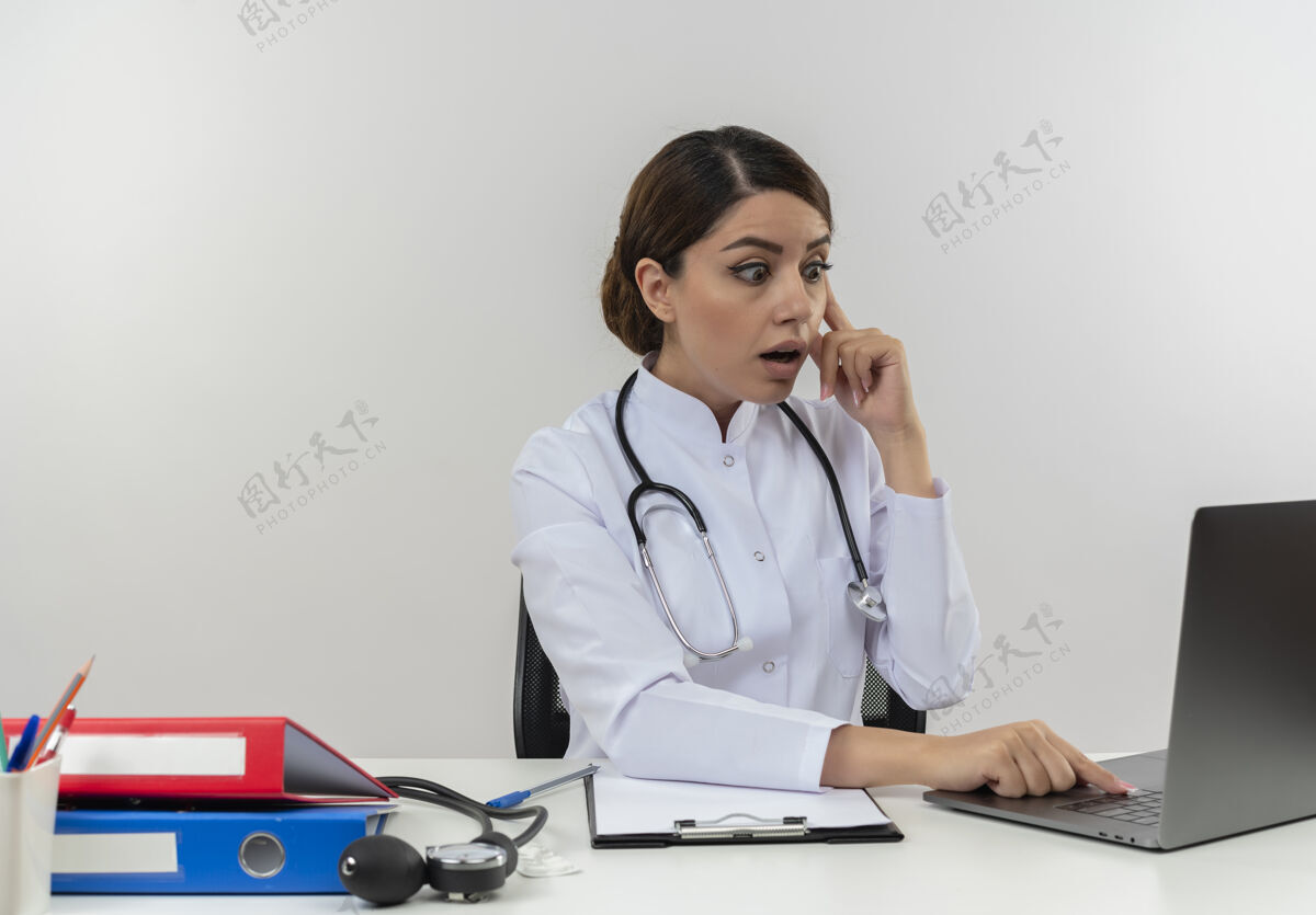 触摸令人印象深刻的年轻女医生穿着医用长袍和听诊器坐在办公桌旁使用医疗工具 看着笔记本电脑触摸头部与手指隔离印象个人坐着