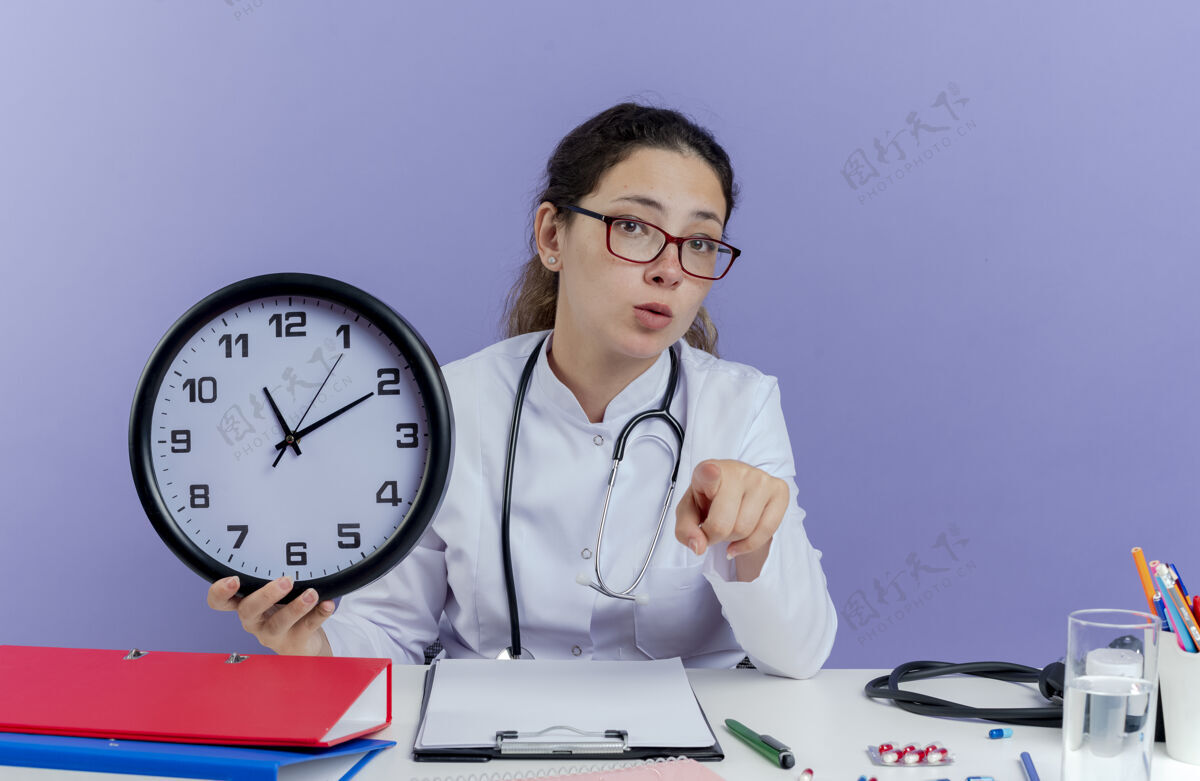穿着令人印象深刻的年轻女医生穿着医用长袍和听诊器坐在办公桌前 手持医疗工具 看着时钟 指着孤立的长袍成人坐着