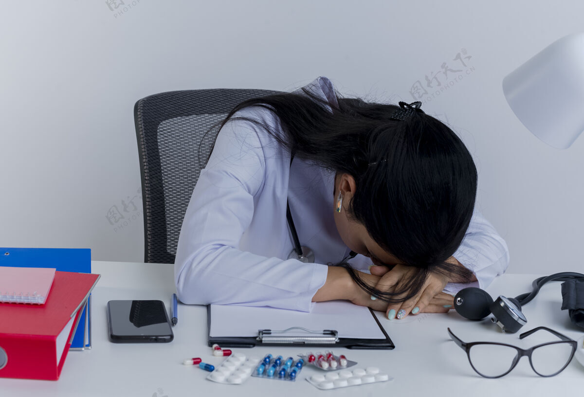 医生疲惫的年轻女医生穿着医用长袍和听诊器坐在办公桌旁 手放在办公桌上 头放在手上累了女人人