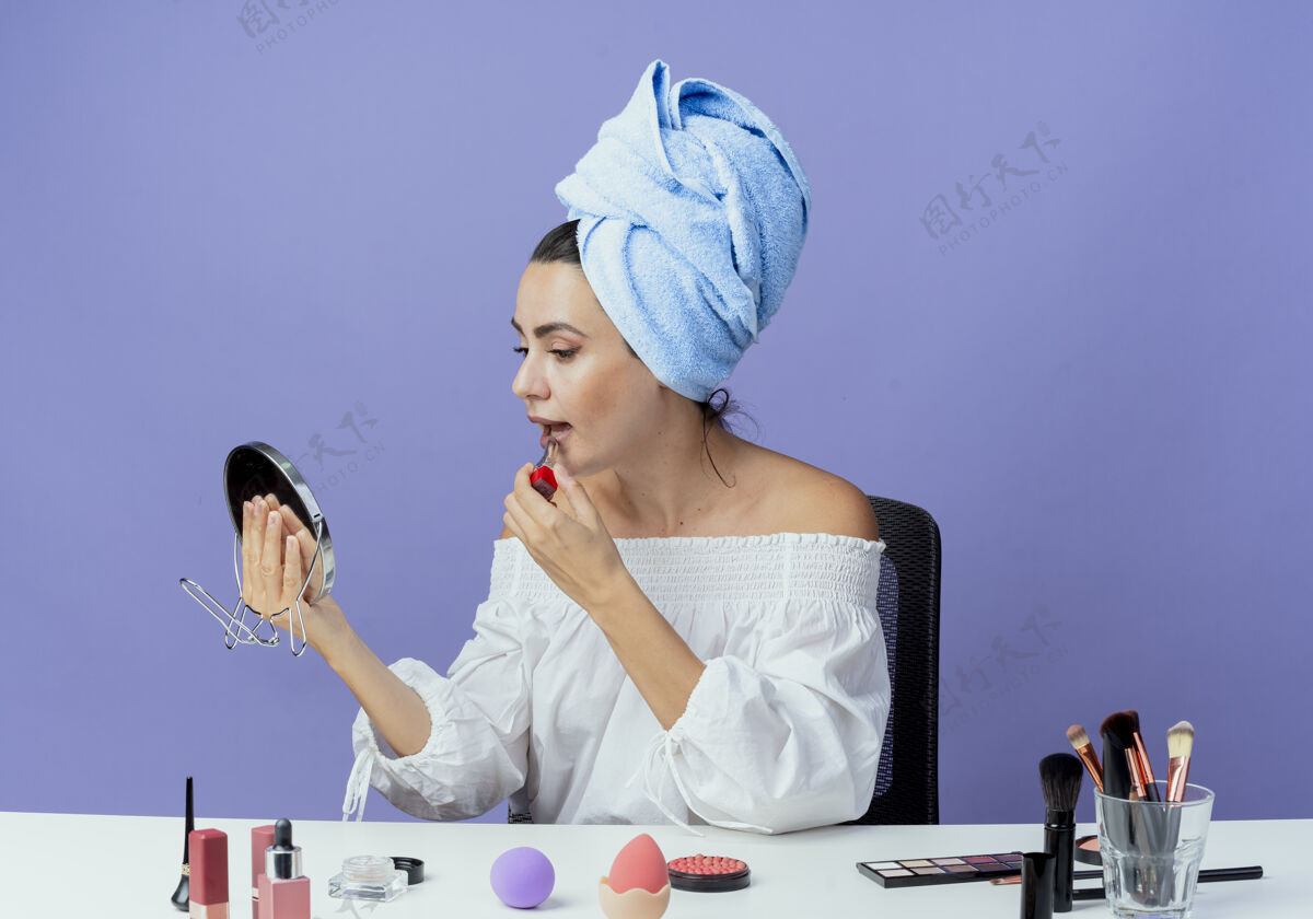 漂亮可爱的漂亮女孩裹着发巾坐在桌子上 拿着化妆工具拿着唇膏 看着镜子在紫墙上孤立口红毛巾女人